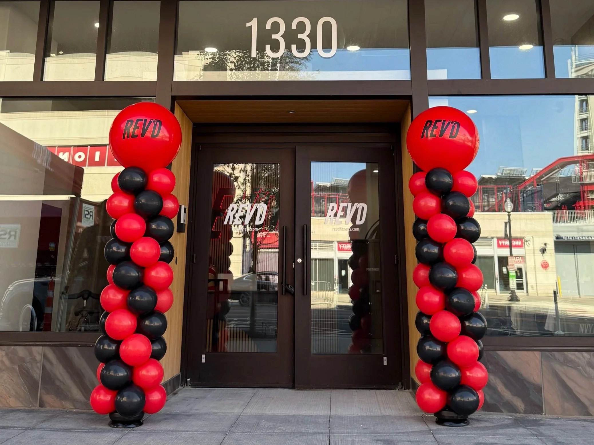 The entrance of a building decorated with red and black balloon columns on both sides of the door with the word "REVD" on large red balloons at the top of each column. Balloon install for grand opening REVD fitness in Navy Yard Washington DC