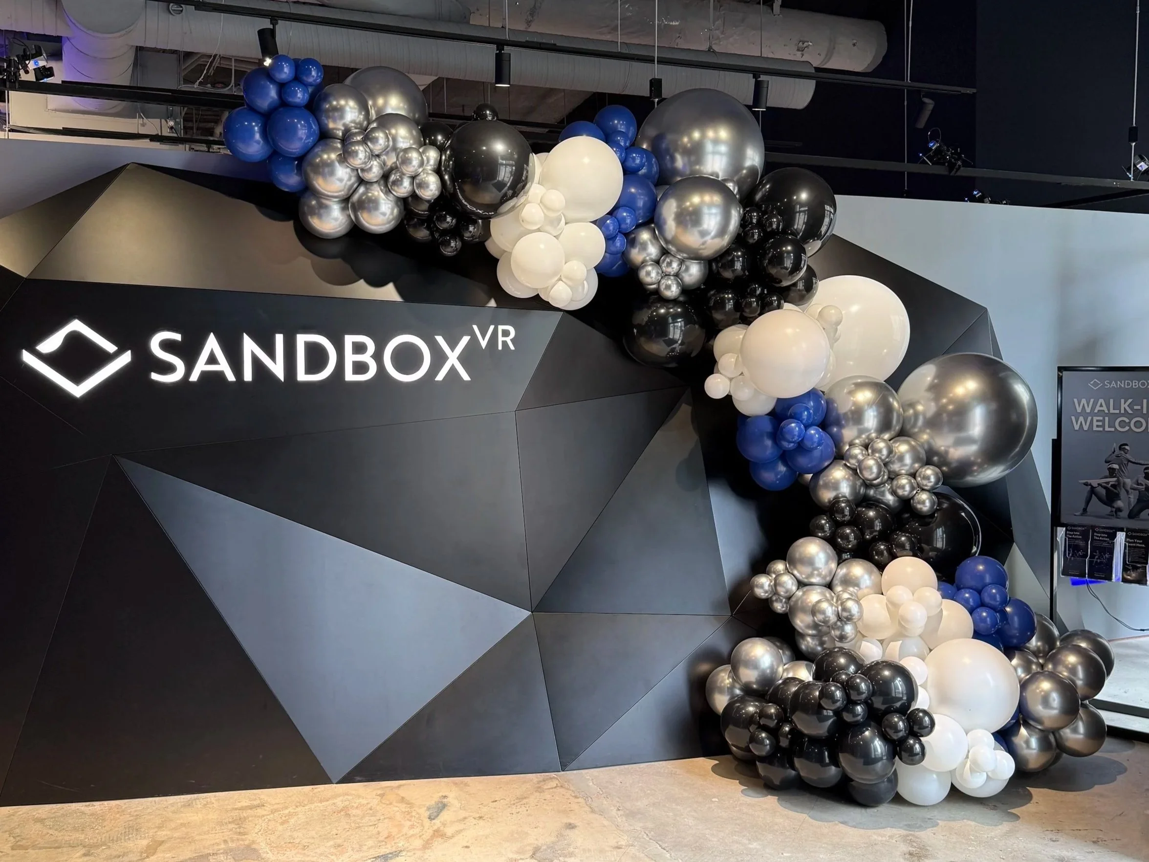 Balloon half arch with black, white, silver, and blue balloons arranged on a black geometric wall with the logo and text 'SANDBOX VR' and part of an event space. Balloon half arch for grand-opening of Sandbox VR in Baltimore, MD
