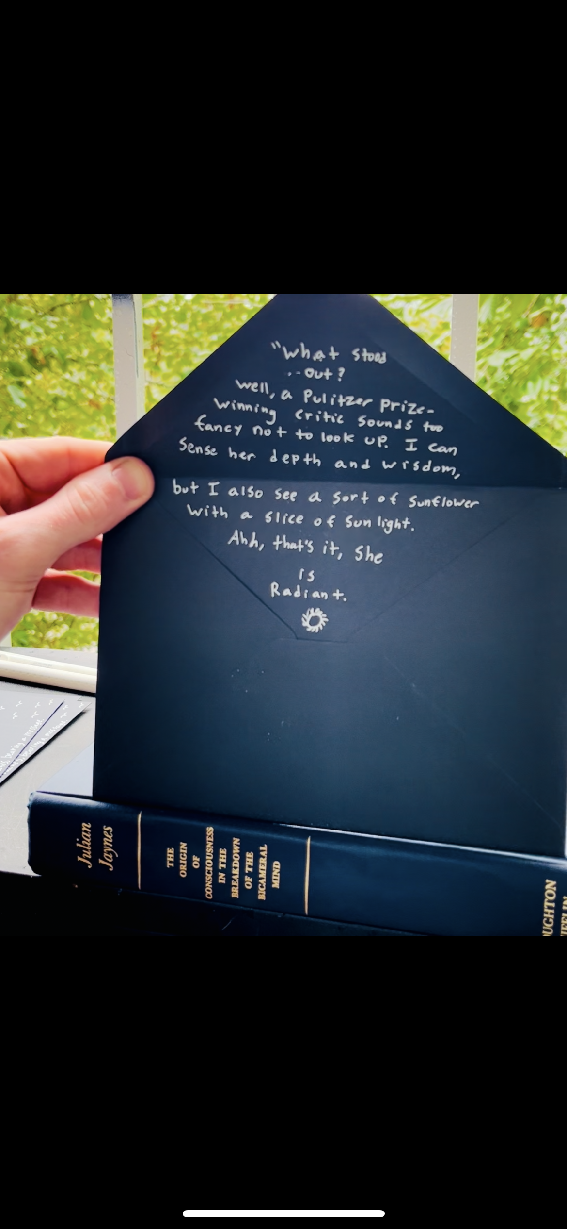 A hand holding a navy blue envelope with a handwritten note inside. The envelope is propped against a book titled 'The Origin of Consciousness in the Breakdown of the Bicameral Mind' by Julian Jaynes. The background features green foliage and a white railing.