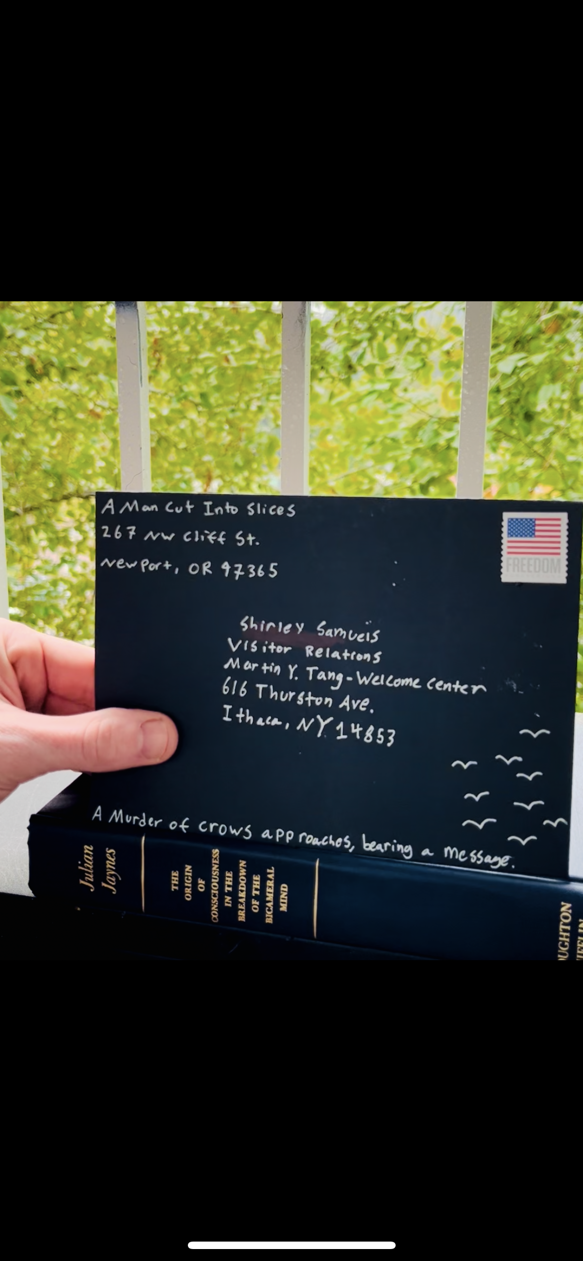 A person holding a black notebook with white handwritten text in front of a green leafy background. The notebook has a small American flag sticker on the top right corner. The handwritten text includes an address in Newport, Oregon, and mentions Shirley Samuels and Martin Y. Tang. The bottom of the page reads "A Murder of Crows approaches, bearing a message." The title of a book by Julian Jones is visible at the bottom.