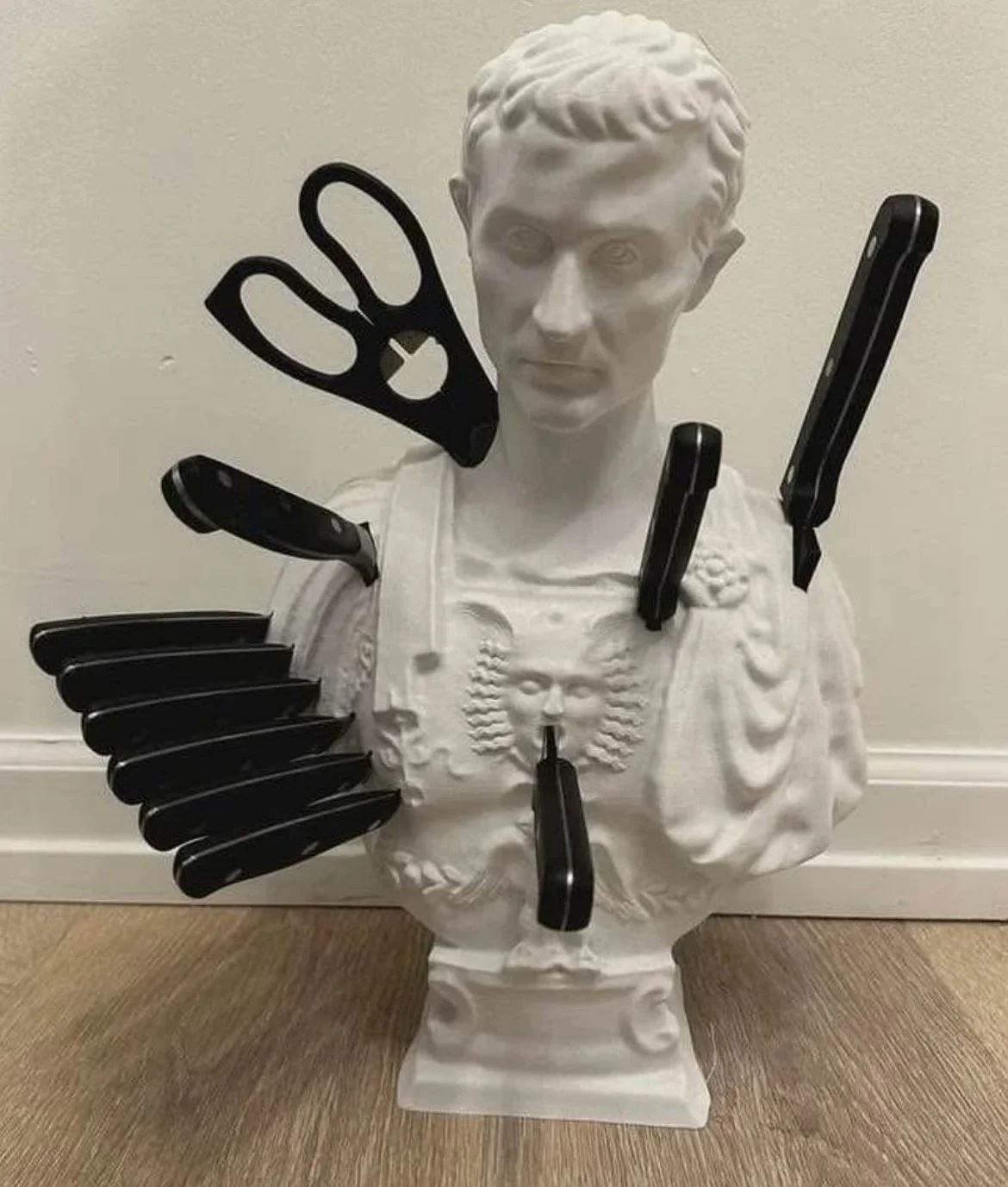Bust of a person with various knives, a pair of scissors, and a small figurine placed on it to create a humorous face.