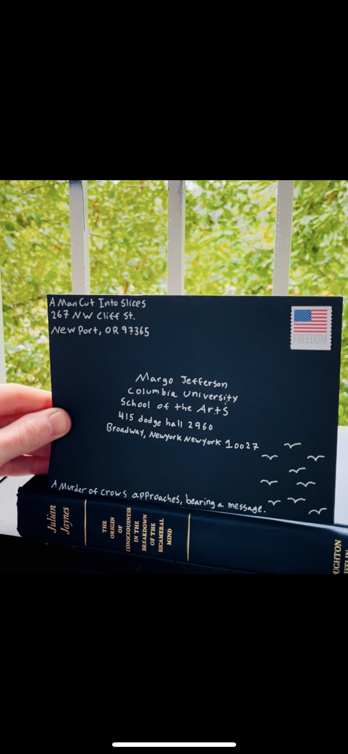A hand holding a navy blue greeting card with handwritten address and a stamp with an American flag and the word 'FREEDOM.' Below the card, a hardcover book titled 'THE ORIGIN OF CONSCIOUSNESS IN THE BREAKDOWN OF THE BRAIN' by Julian Jaynes is visible. The background shows green foliage and a white fence.