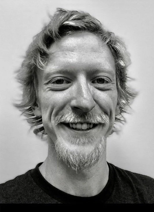 Photograph of a man with light-colored hair, a beard, and a mustache, smiling at the camera, wearing a dark shirt.