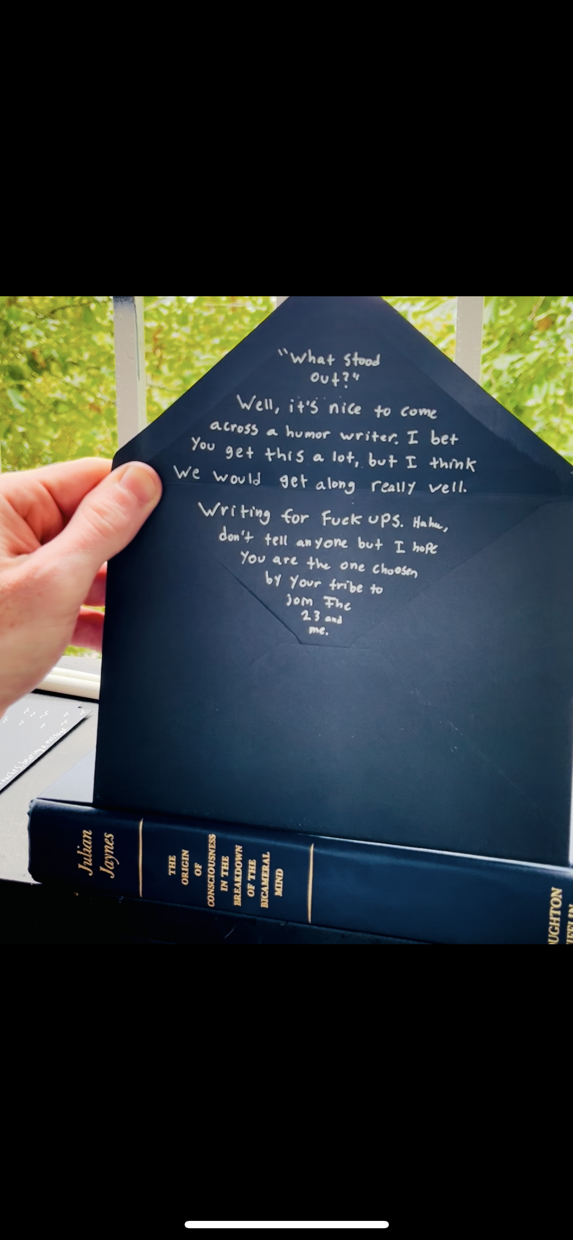 A hand holding a navy blue envelope with a handwritten note inside, written in white ink. The note discusses humor writing and personal preferences. The envelope is on a table with a large navy blue book titled 'The Origin of the Consciousness in the Breakdown of the Brain of the Mind' by Jablian Joynes underneath. A window with green foliage can be seen in the background.