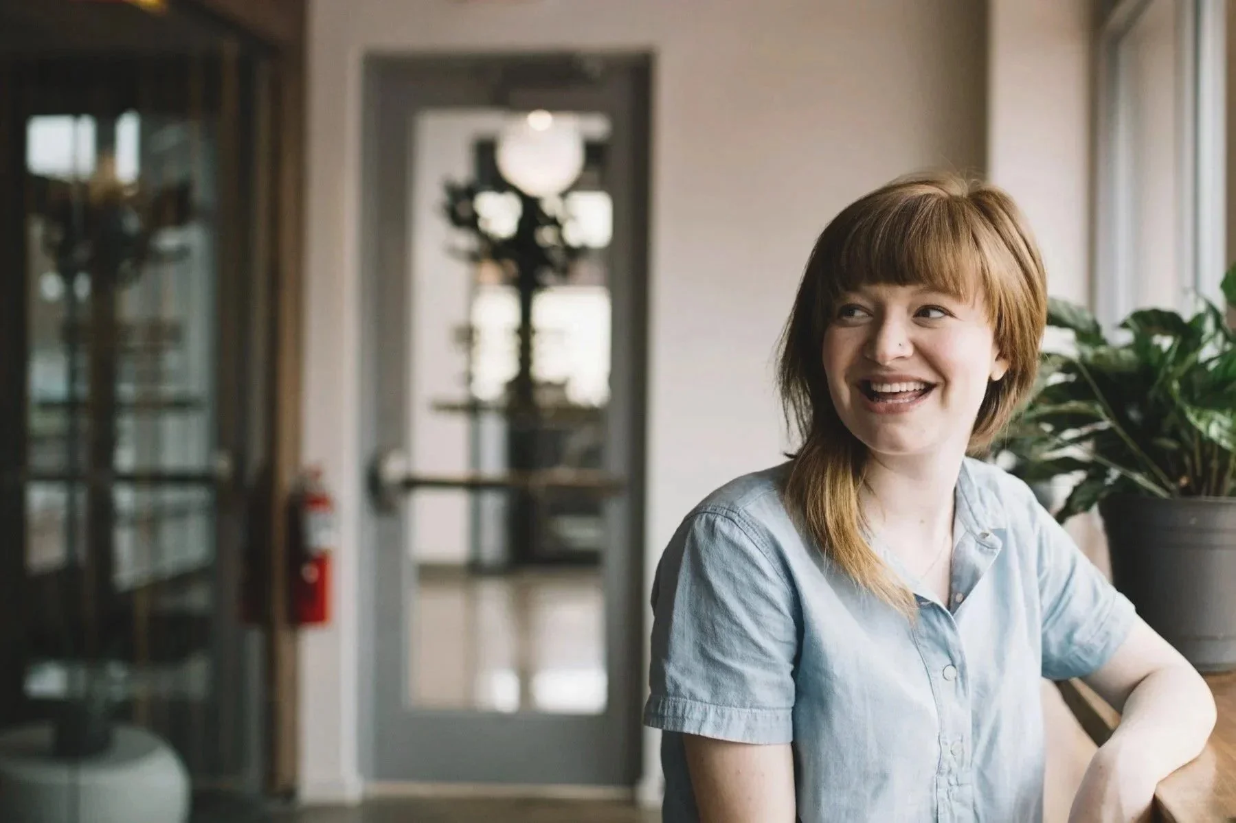 A woman with red hair smiling while sitting at a table in a bright room with large windows, plants, and a blurred background.