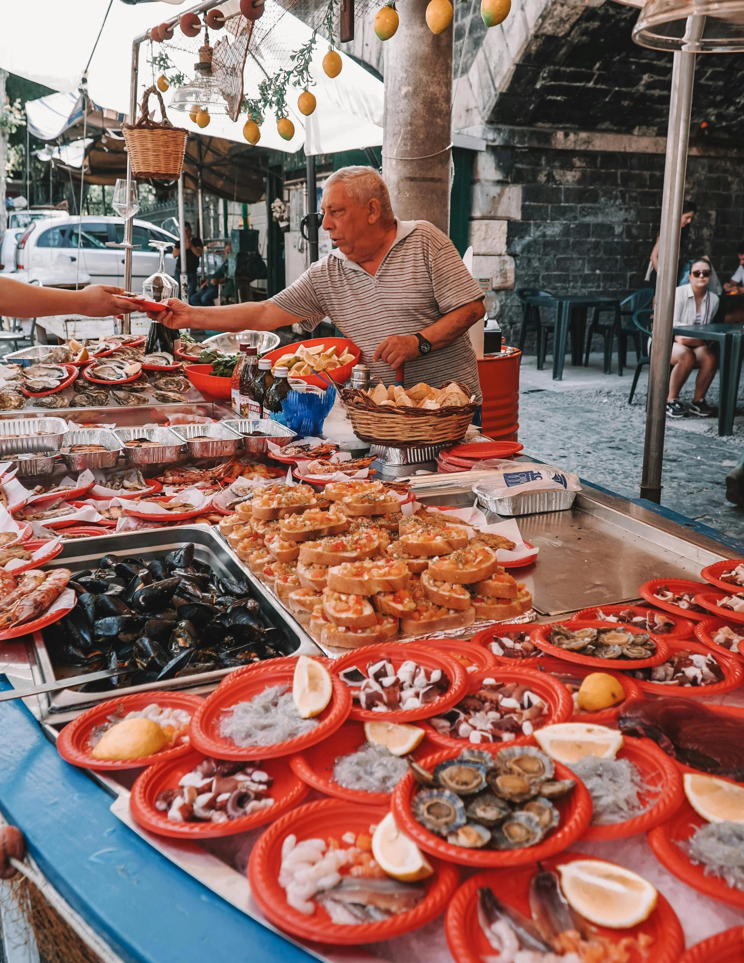 Man at outdoor seafood market with various shellfish, oysters, lemon wedges, and seafood dishes on red plates.