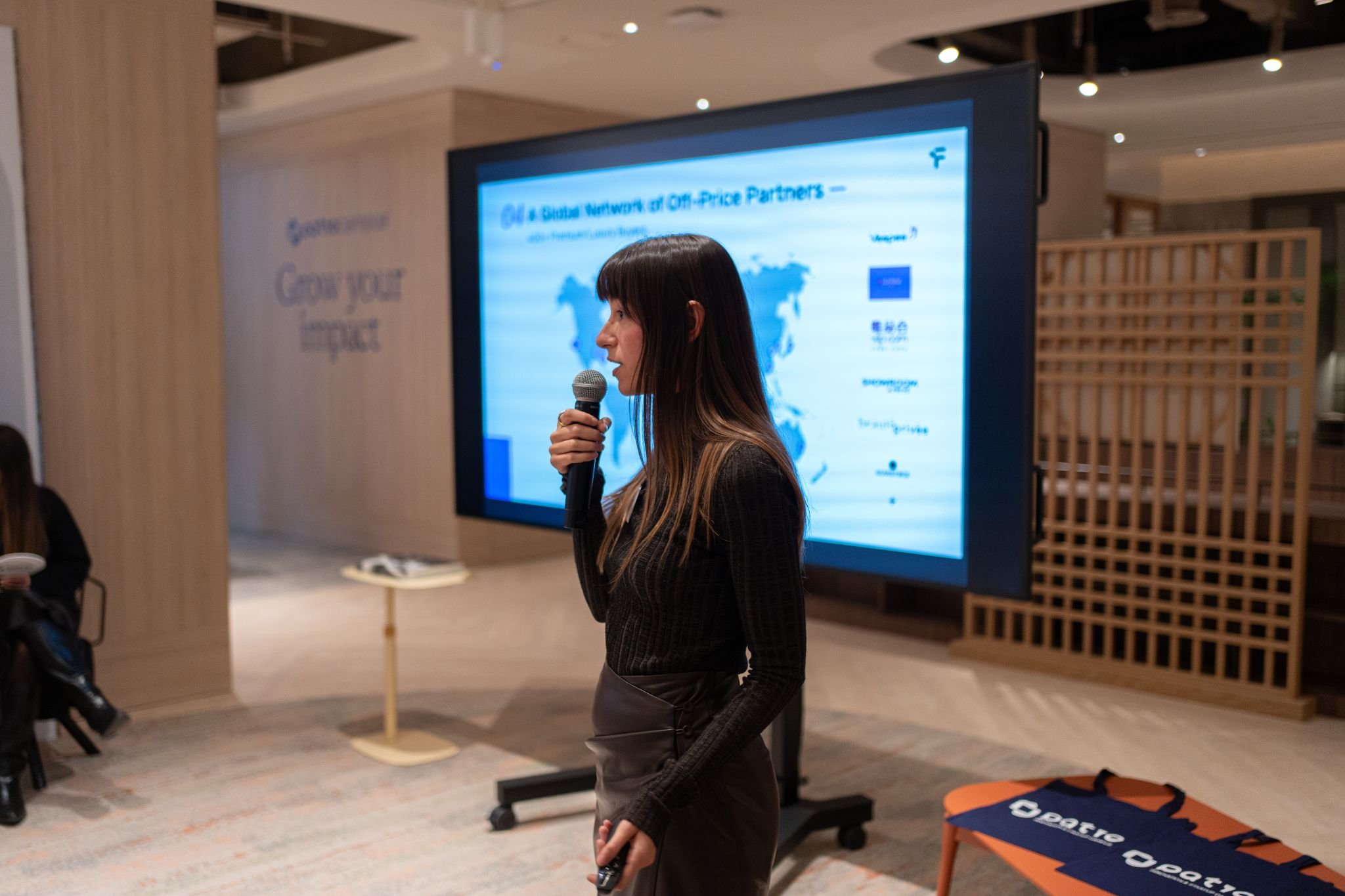 A woman holding a microphone and speaking at a presentation or conference in front of a large screen with a world map and text.
