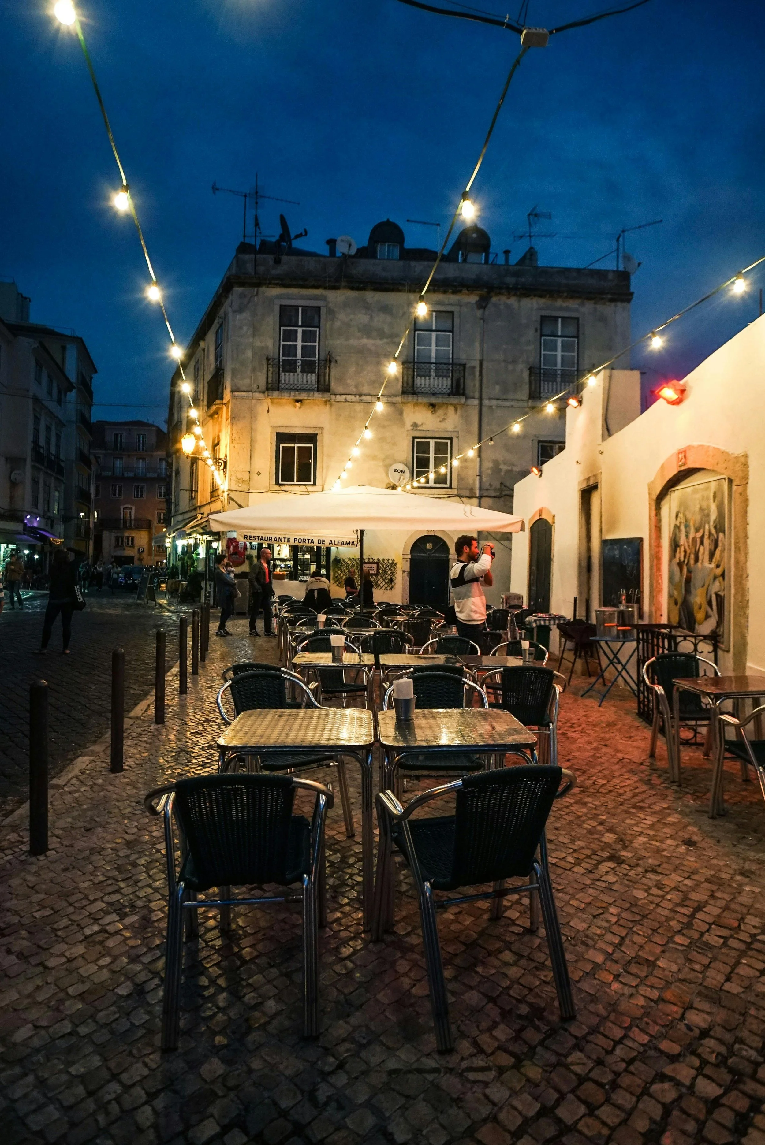 Empty outdoor cafe on cobblestone street during evening with string lights overhead, a man taking a photo, and a historic building in the background.
