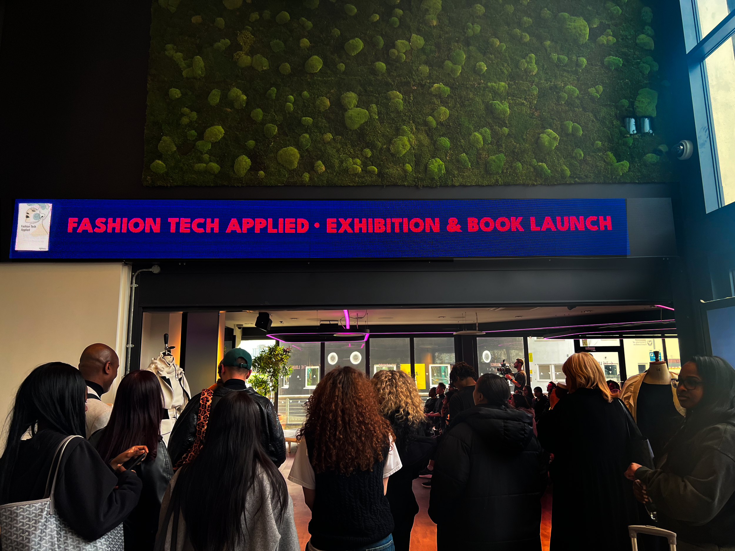 Crowd of people waiting indoors for the Fashion Tech Applied exhibition and book launch, with green moss-like wall art and a digital display with event information.