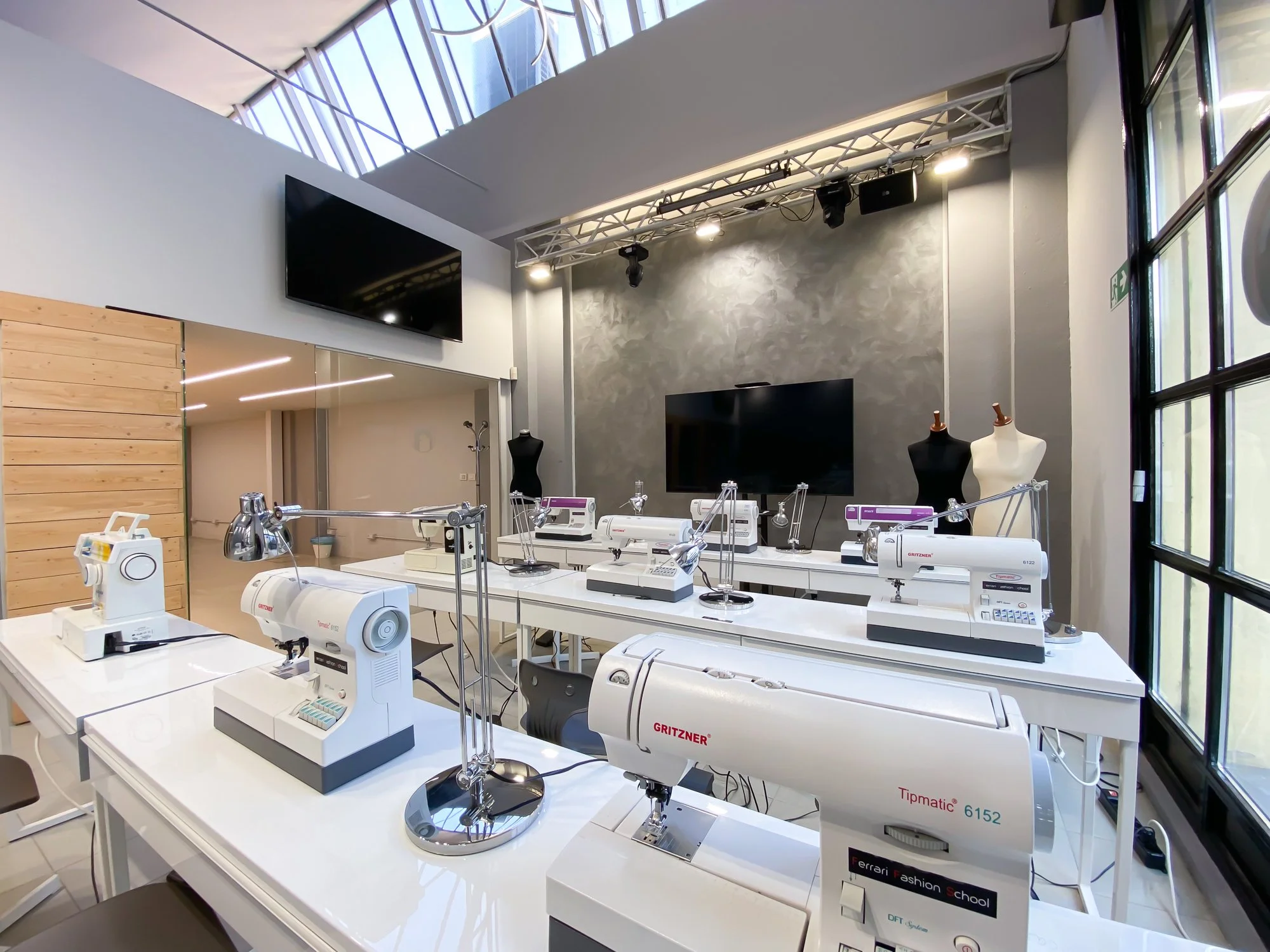 A sewing class or workshop with multiple sewing machines on white tables, mannequin torsos, and large monitors on the walls in a modern, well-lit space.