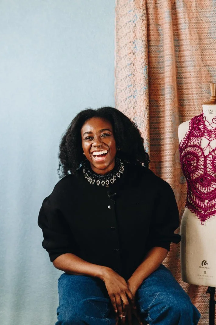 A woman with curly black hair smiling and laughing, sitting in front of a sewing mannequin with purple lace fabric, and a pastel-colored curtain in the background.