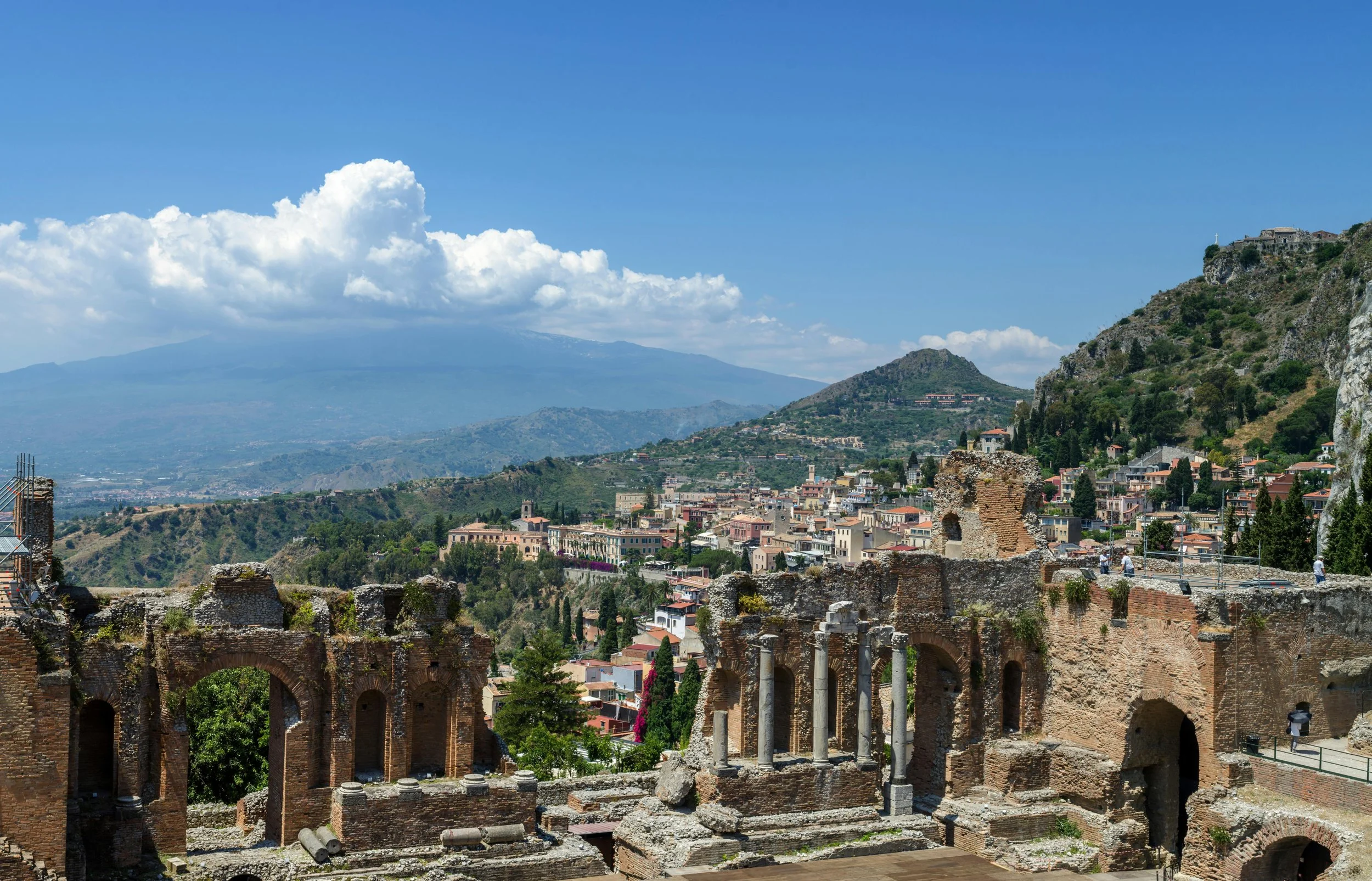 Ancient ruins with arches and columns, overlooking a hillside city with colorful buildings, green trees, and a mountain in the background under a blue sky with clouds.