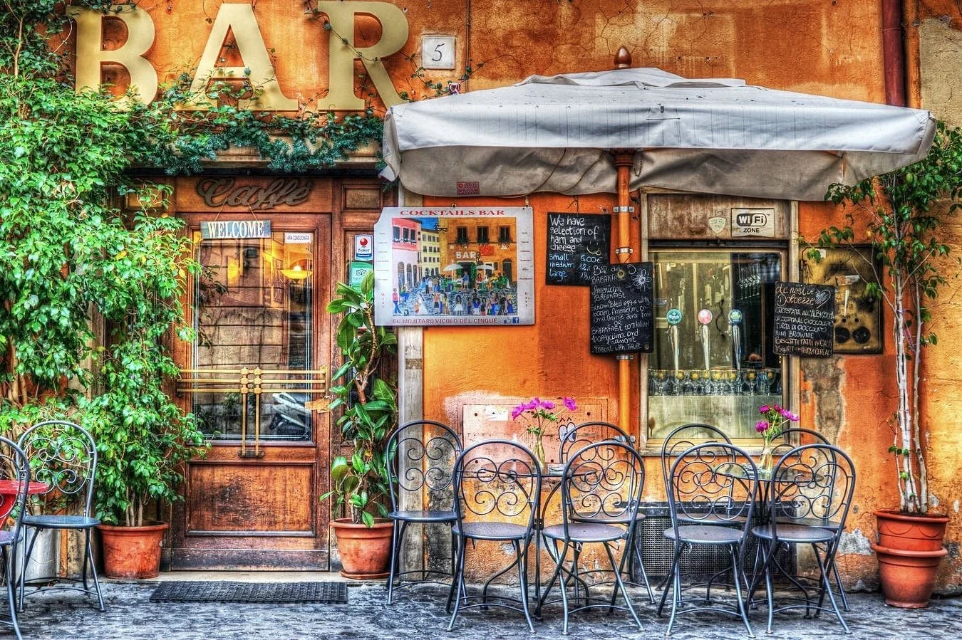 Outdoor seating area of a small restaurant or café with black wrought iron chairs and small tables with pink flowers in a clear glass vase, green plants in pots, and a large white umbrella. The background features an orange wall with signs, blackboards with menu items, a wooden door with glass panels, and a window with a sign indicating Wi-Fi zone.