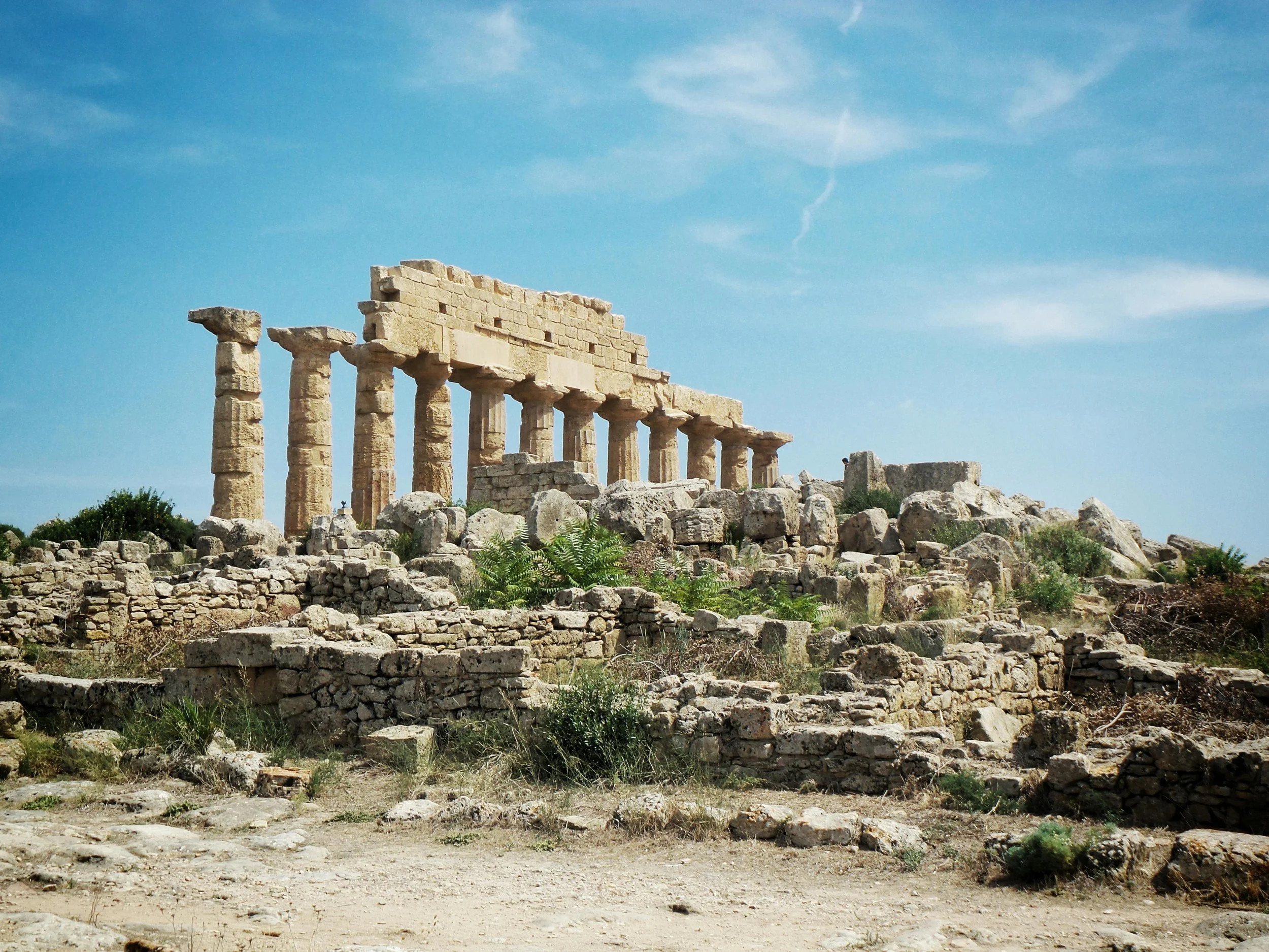 Ancient Greek ruins with several stone columns and a structure on top, surrounded by rocks and vegetation under a blue sky.