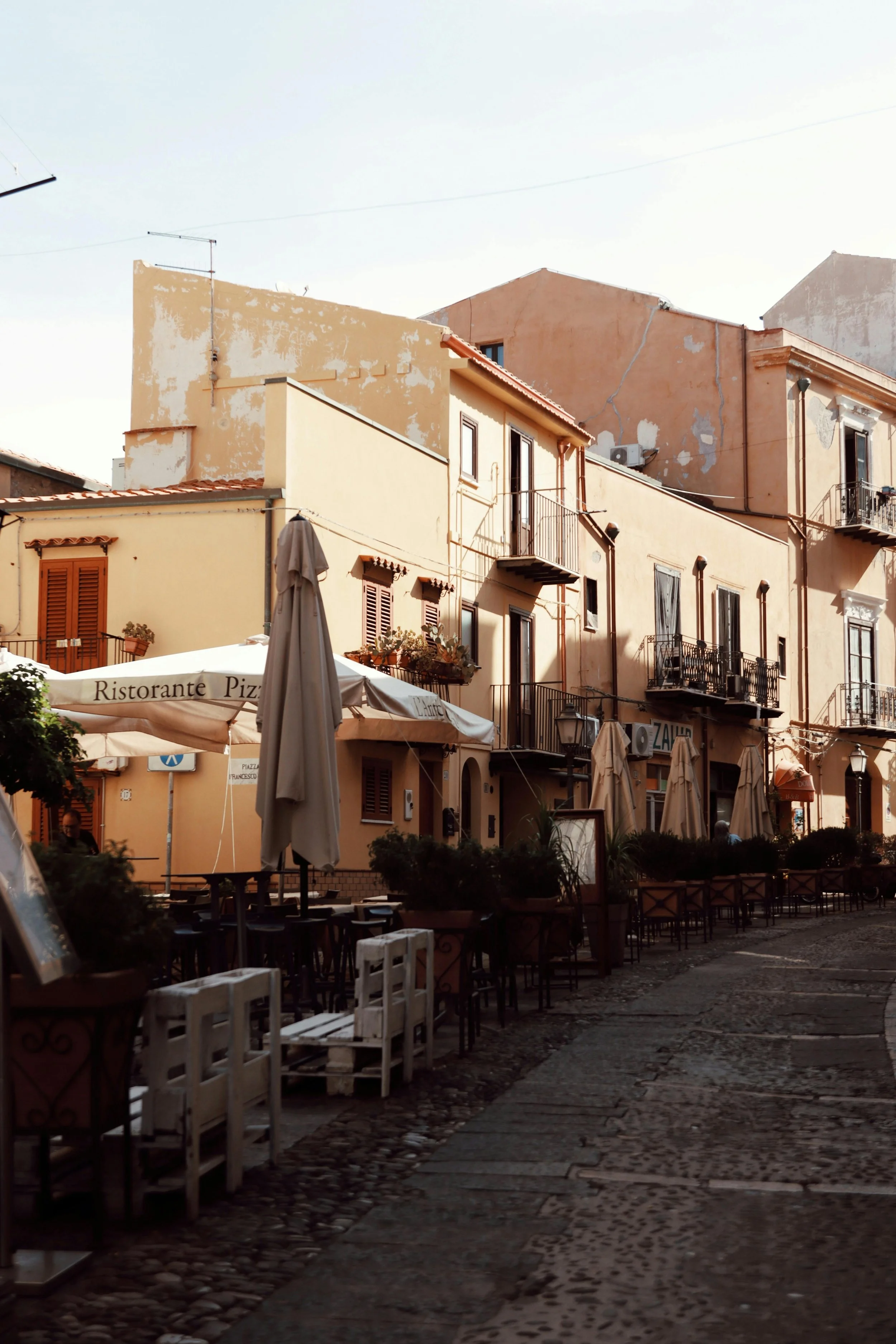 A cobblestone street with outdoor tables and umbrellas, European-style buildings with balconies, and a clear sky.