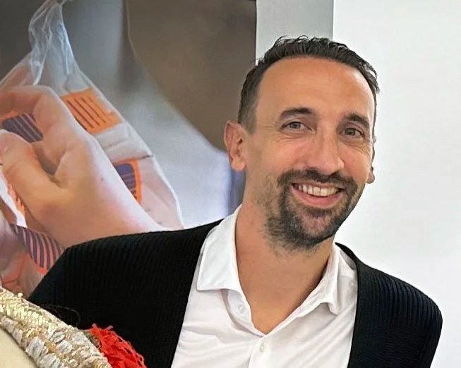 Smiling man with short dark hair and a beard, wearing a white shirt and black blazer, standing indoors with a blurred medical image in the background.