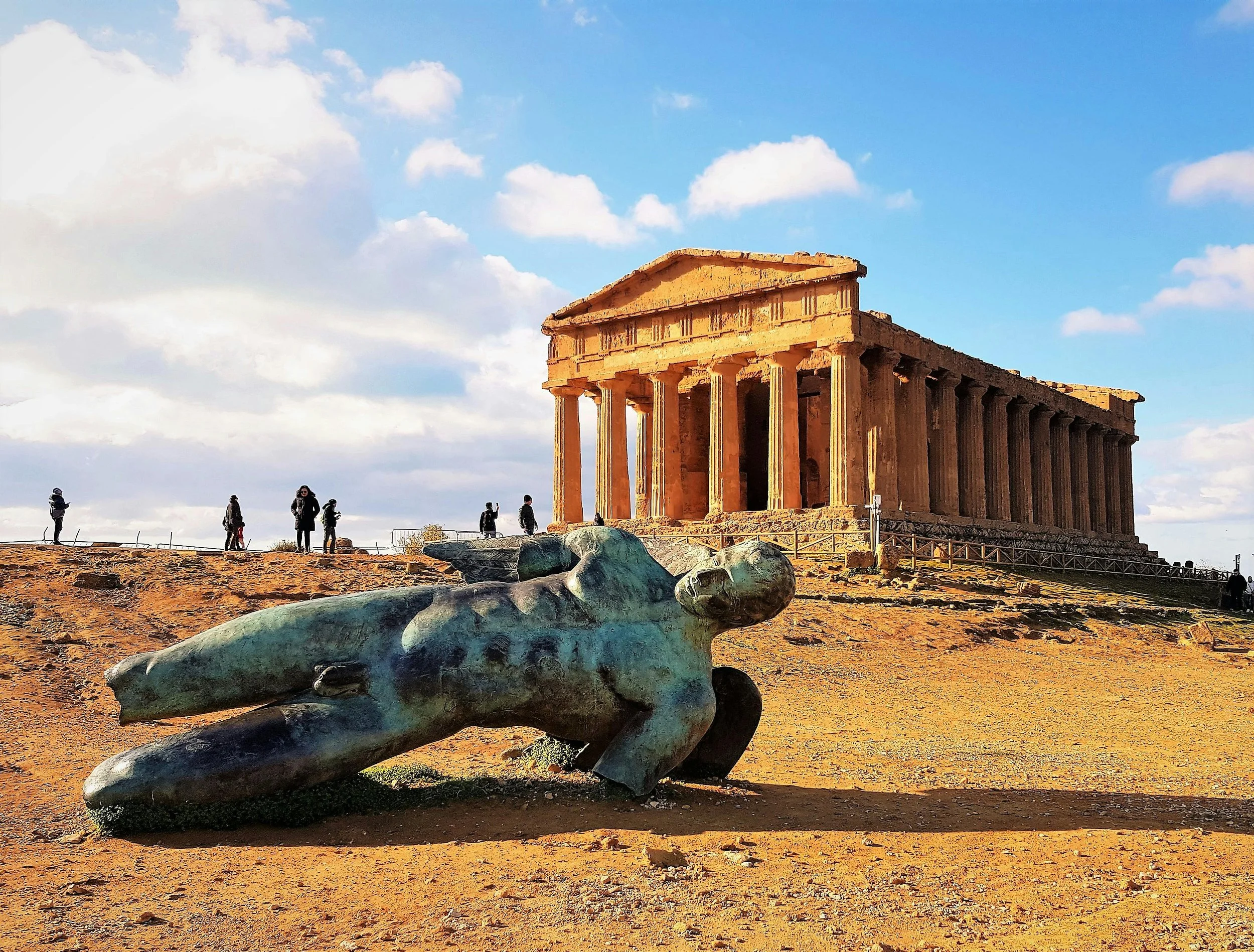 Ancient Greek temple with tall columns, situated on a hill, with visitors walking around, and a large, fallen bronze statue in the foreground. The sky is partly cloudy.