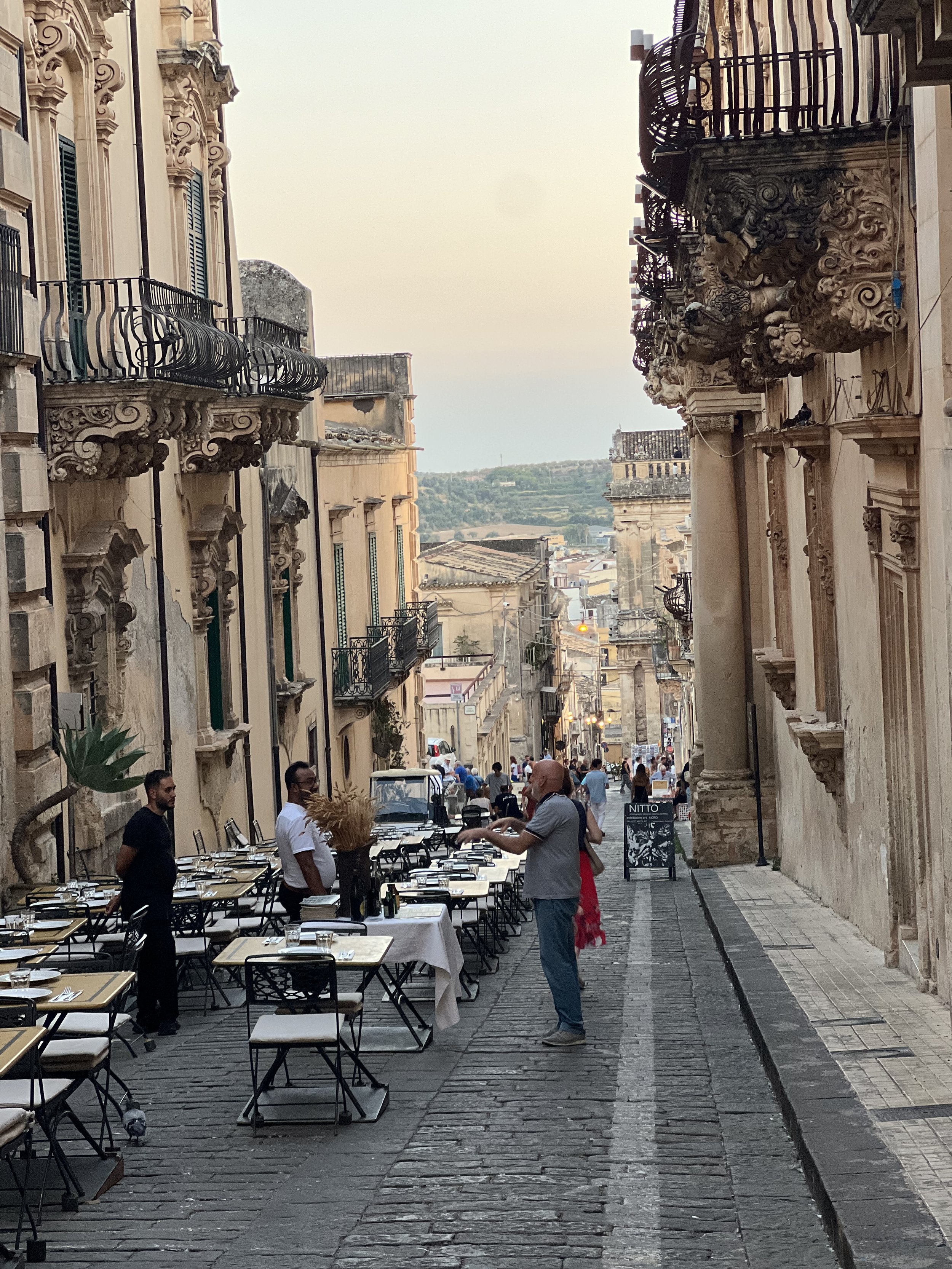Street view with outdoor cafe tables set along a narrow cobblestone sidewalk, surrounded by ornate historic buildings with balconies, in a European city during sunset.