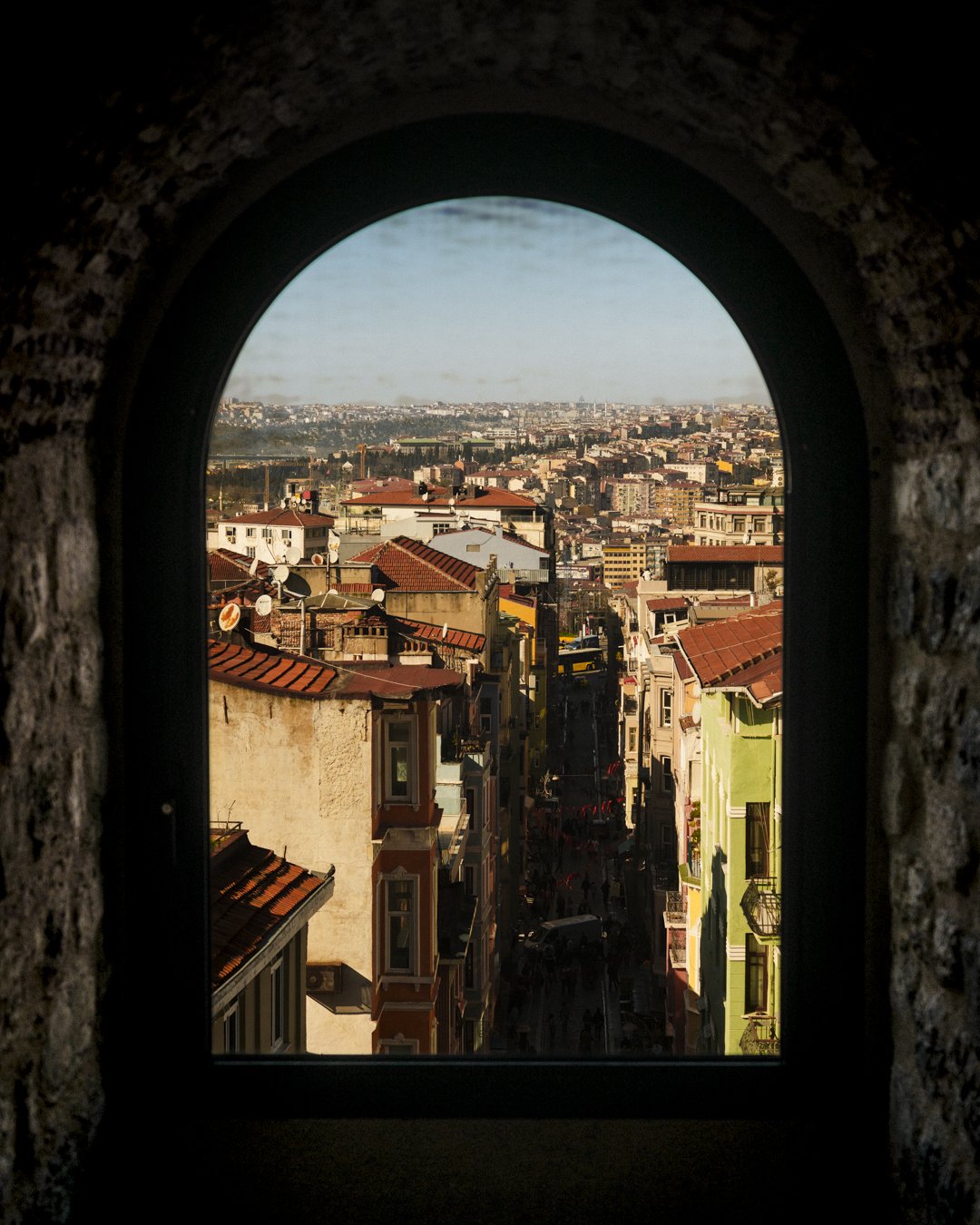View of city streets and rooftops through an arched window, with buildings tightly packed and a distant cityscape visible.