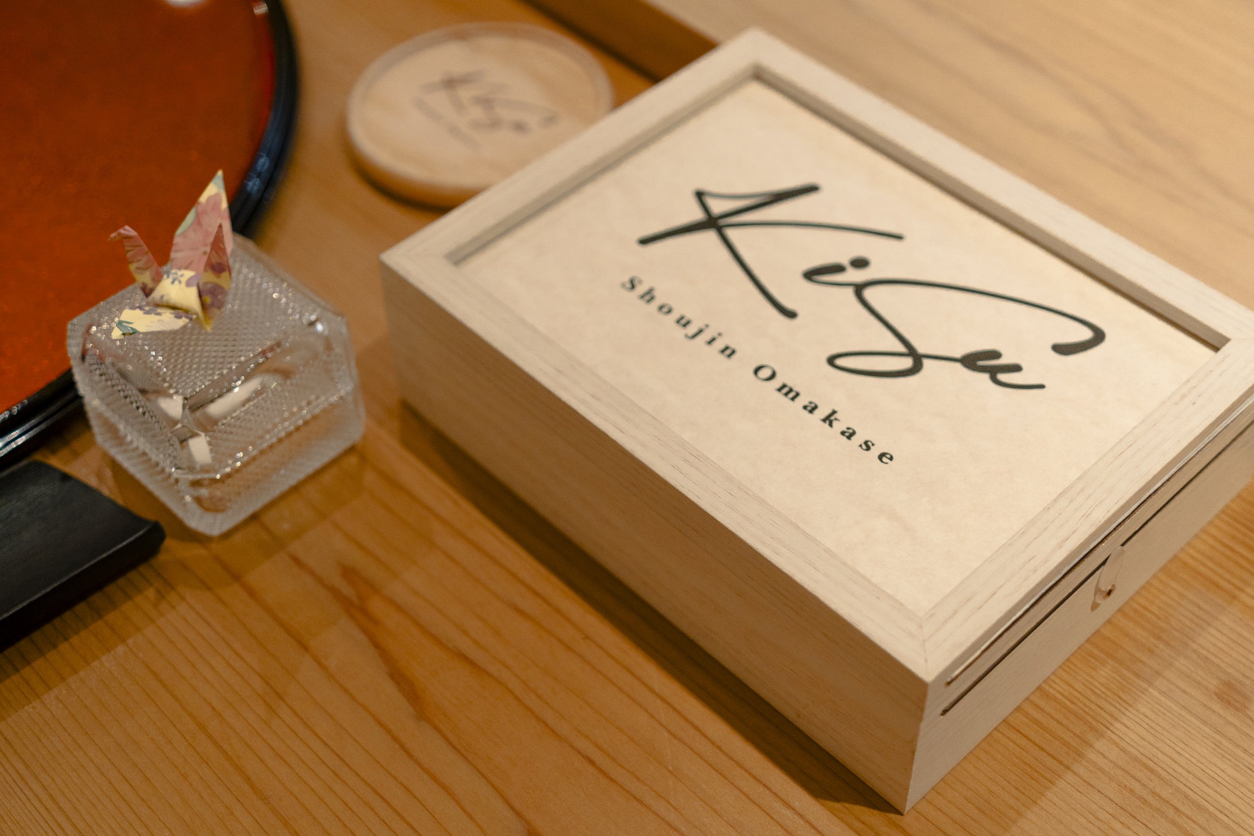 A wooden box labeled "Kisue Shoujin Omakase" on a light-colored wooden table, with a small glass container holding a paper crane next to it. A round wooden coaster is partially visible in the background.