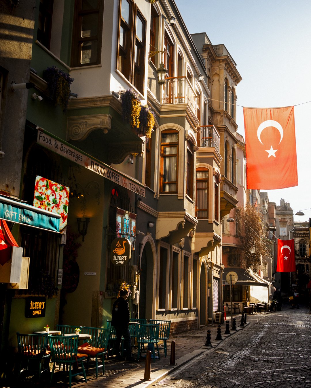 Cobblestone street with colorful buildings, Turkish flags hanging, outdoor café seating, and sunlight casting shadows.