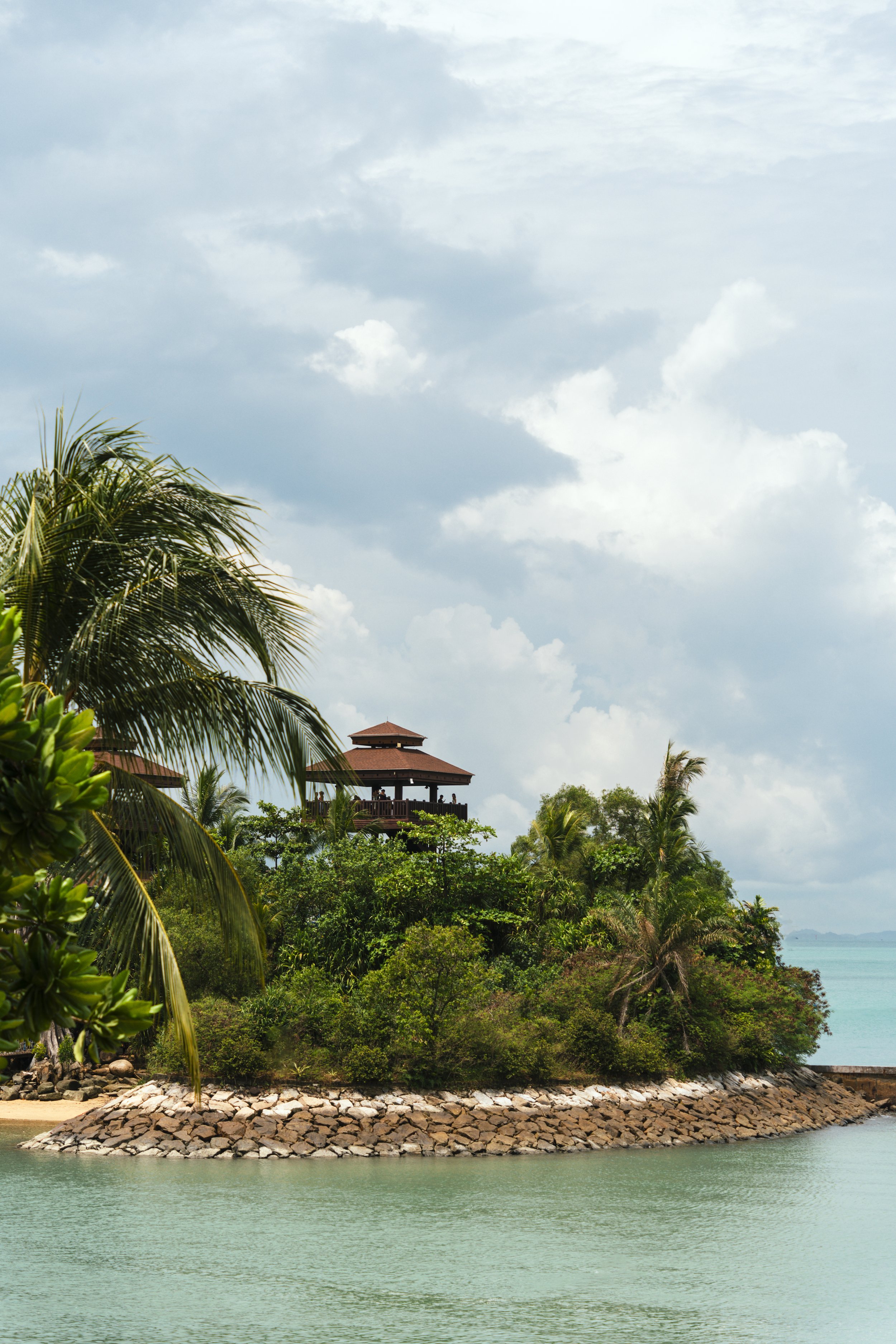Island landscape with tropical trees and a wooden pavilion near a rocky shoreline, under a cloudy sky.