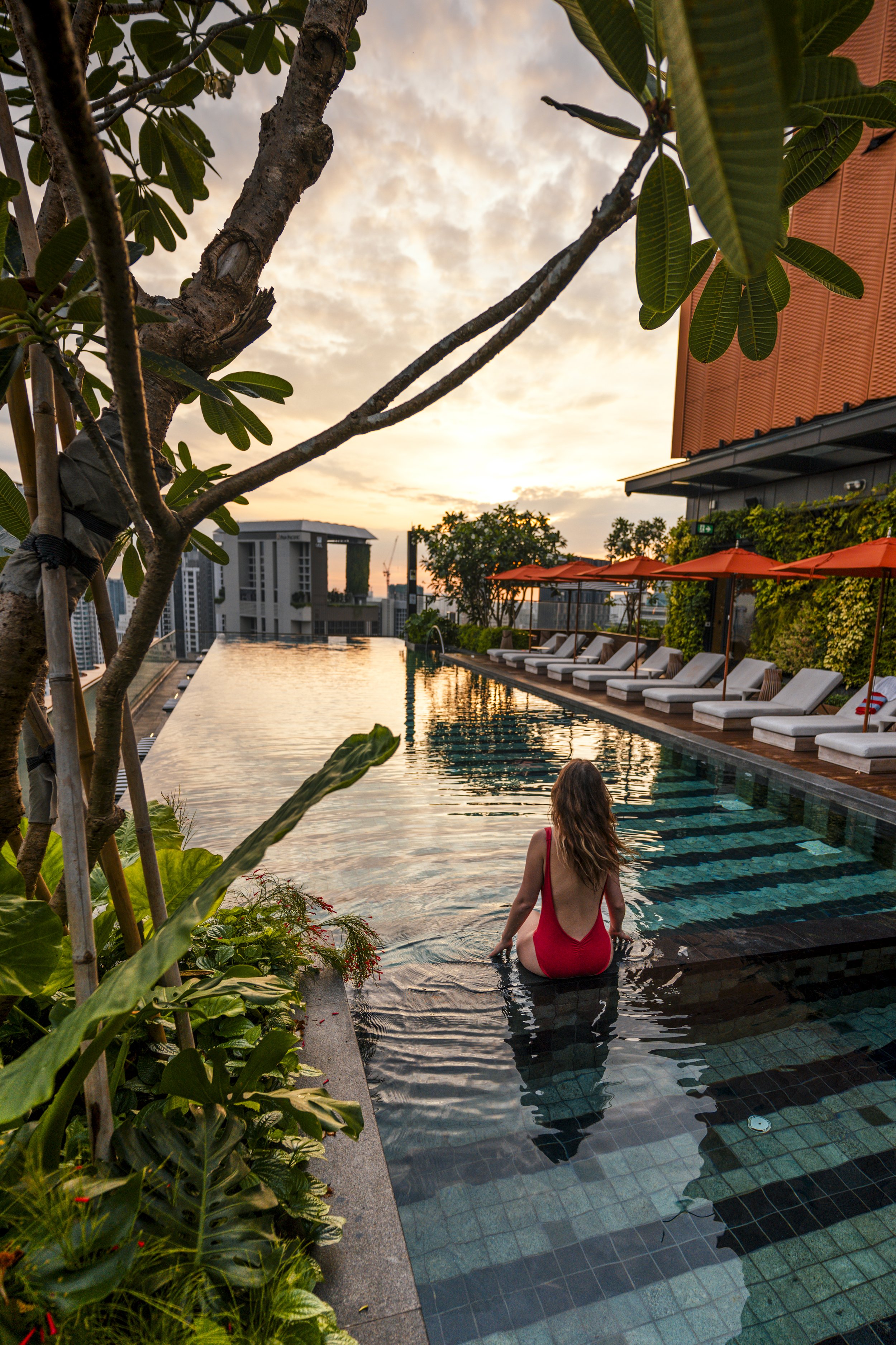 Woman in red swimsuit sitting by an outdoor infinity pool with city skyline at sunset, surrounded by lounge chairs and greenery.