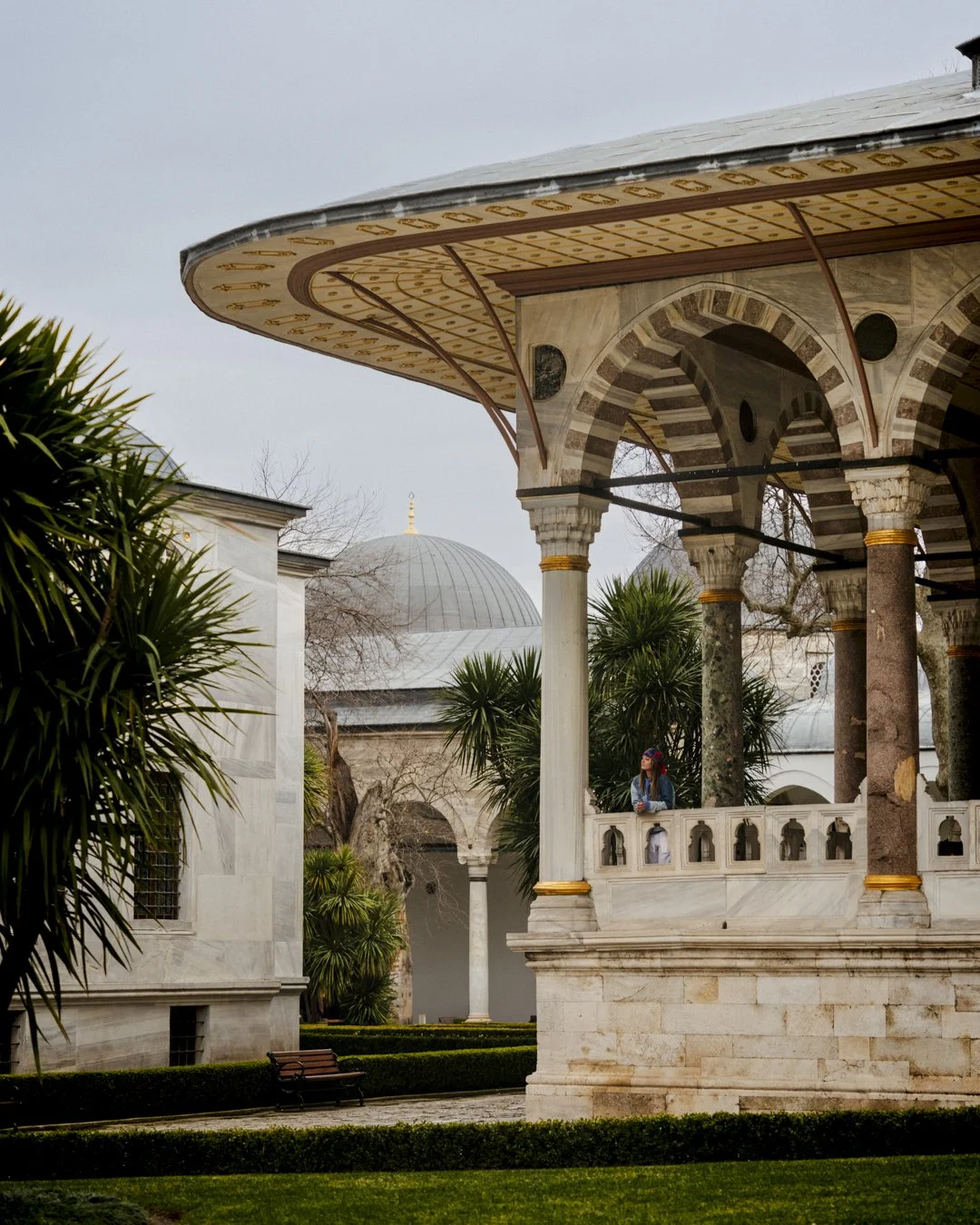 Historic architectural structure with ornate arches and columns, surrounded by lush greenery and a distant dome in a garden setting.
