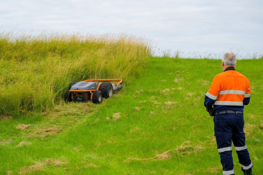 A person in an orange safety jacket and navy pants stands on a grassy hill next to a small autonomous mowing robot that has run off course and is stuck in the grass.
