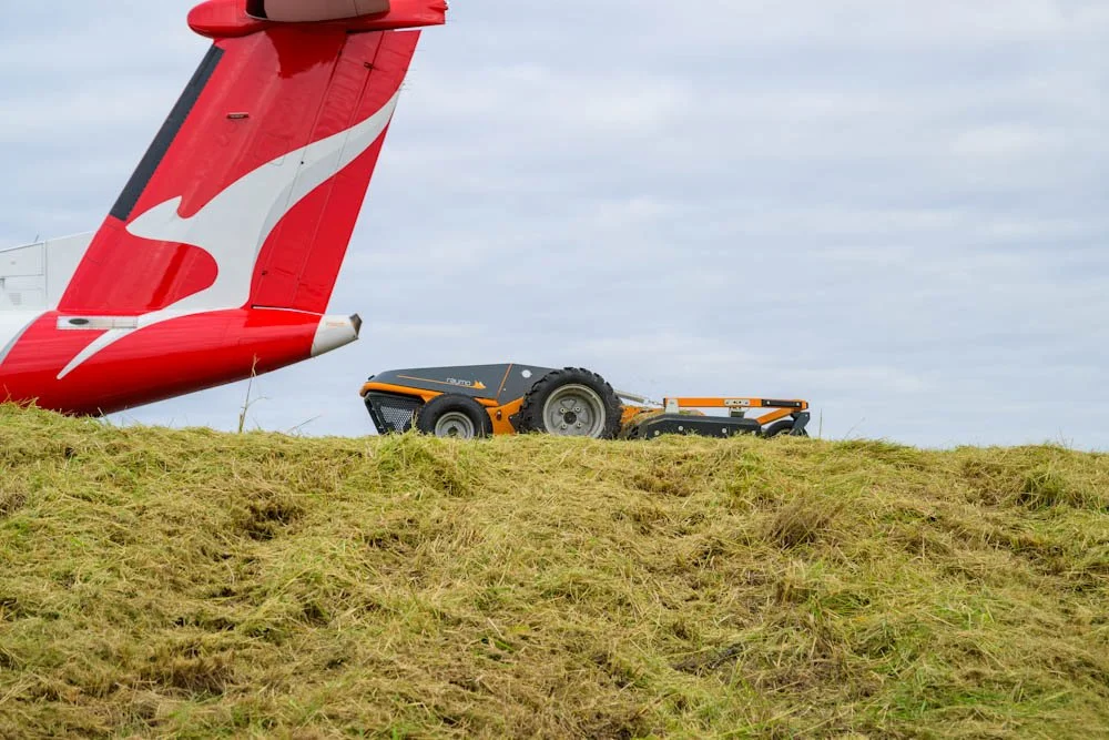 Part of a red airplane's tail in the sky, with a small, orange and black lawn mower on a grassy mound underneath it, and cloudy sky in the background.