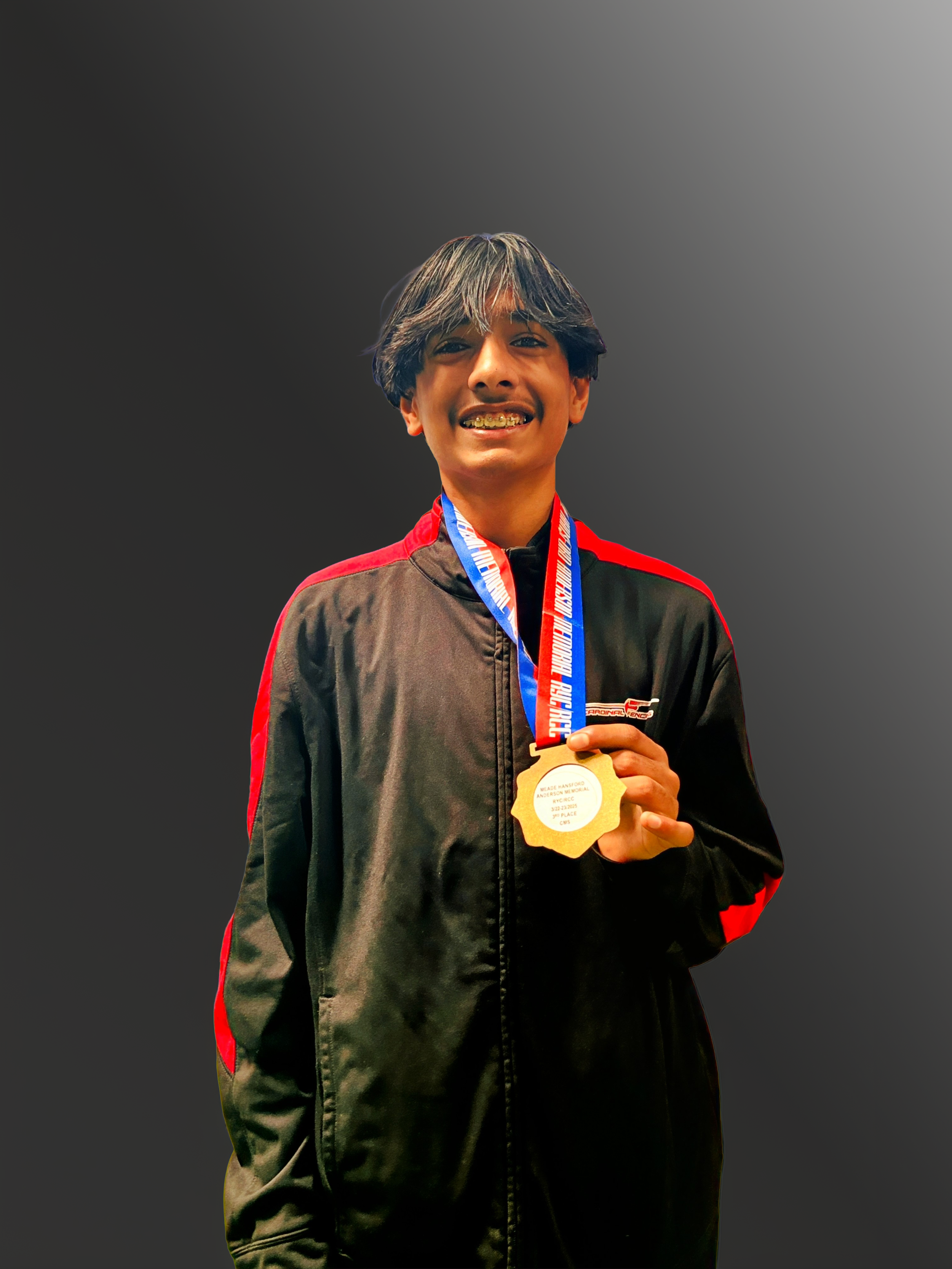 Young boy with dark hair smiling, wearing a black athletic jacket with red accents, holding a gold medal on a blue ribbon.
