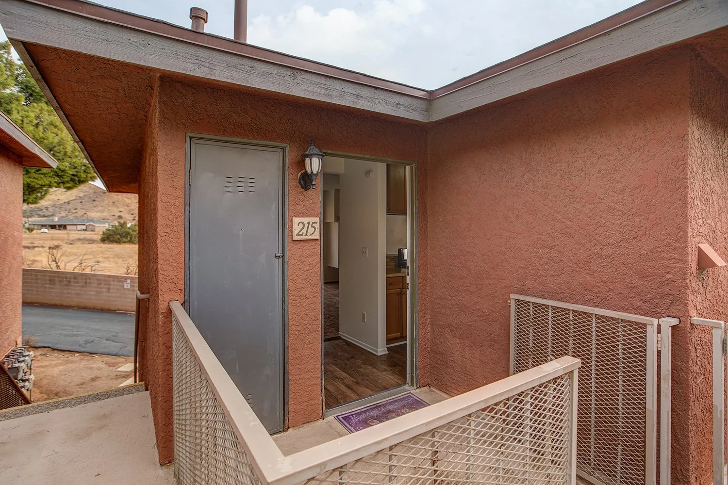This image captures an upper-level apartment entrance, showing an open front door that reveals a glimpse of the interior kitchen alongside an outdoor metal railing.
