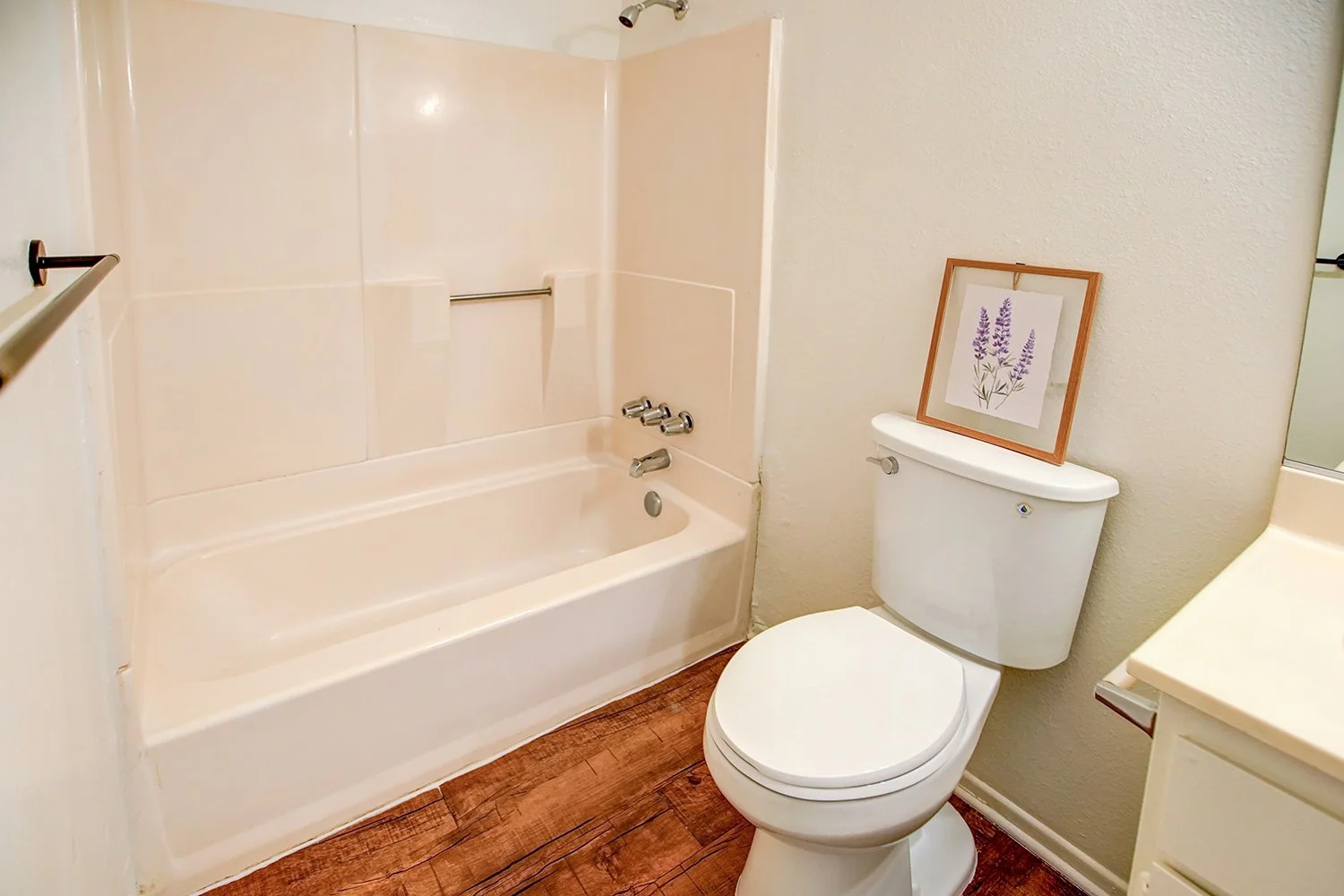 This image shows a compact apartment bathroom featuring wood flooring, a white toilet, and a combined bathtub and shower unit.