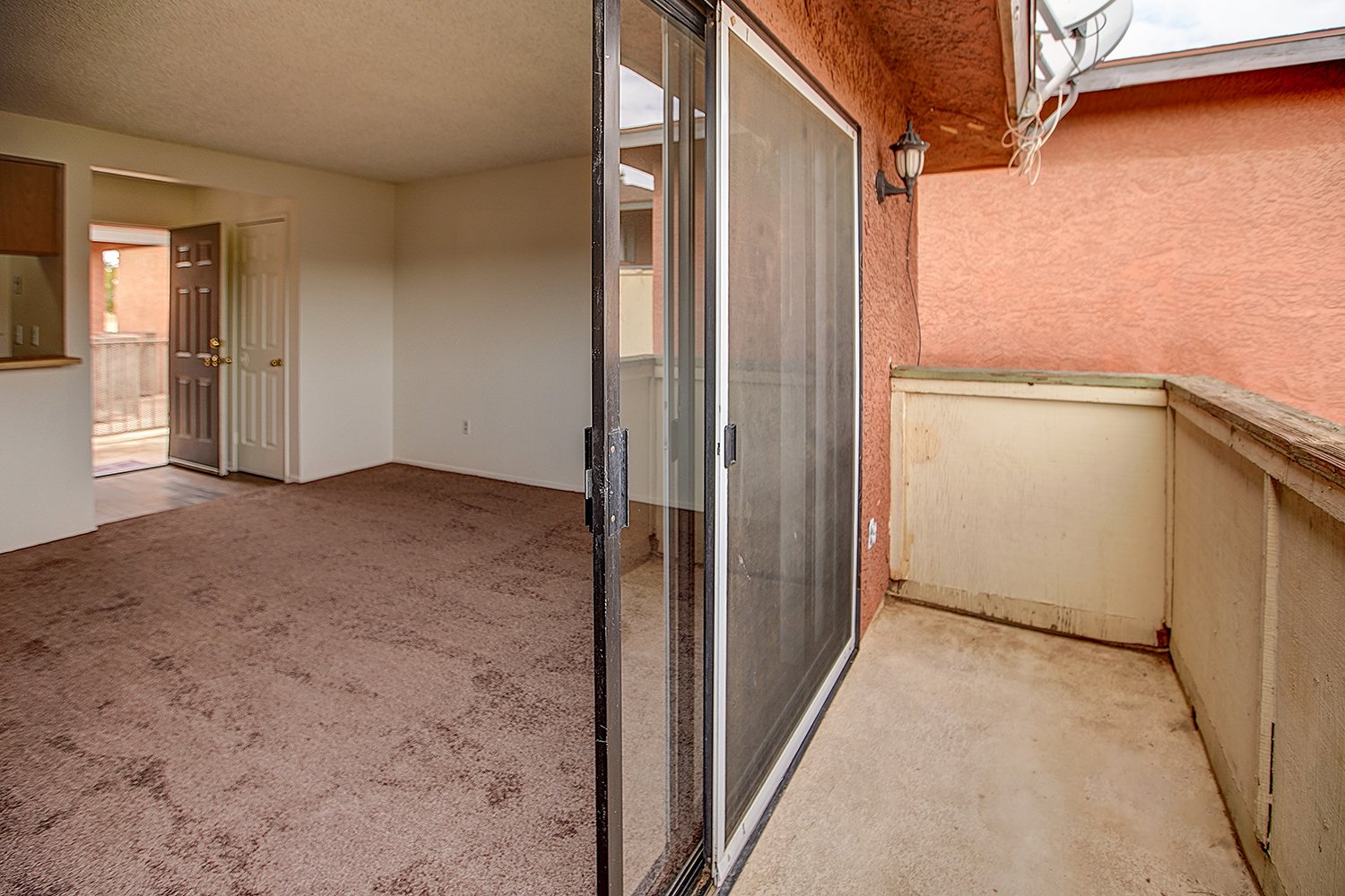 This image shows an outdoor apartment balcony with concrete flooring looking into a carpeted living room through a large sliding glass door.