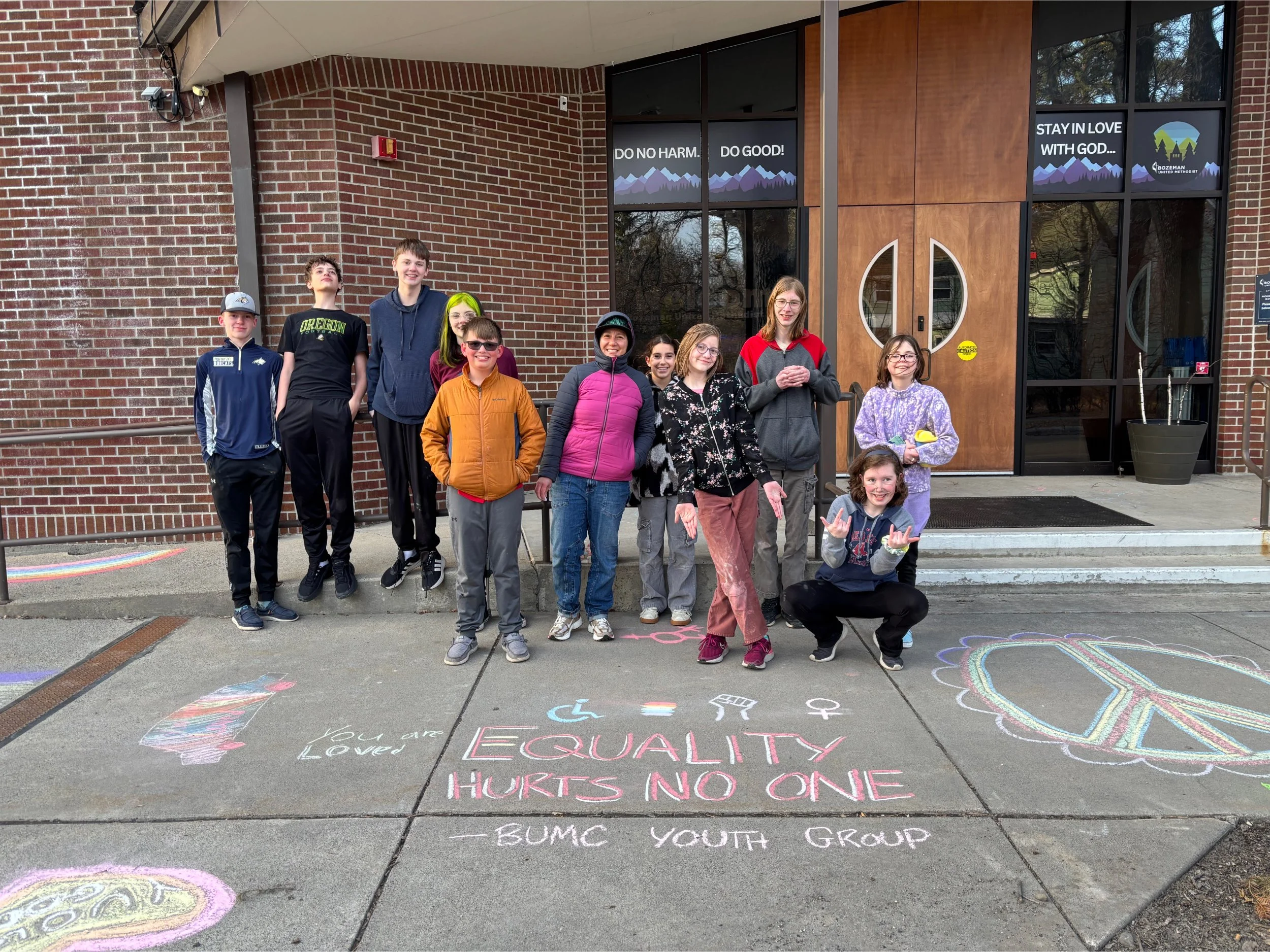 Group of teenagers standing in front of a brick building with chalk drawings and messages on the sidewalk, including a rainbow peace sign and the phrase 'EQUALITY HURTS NO ONE.'