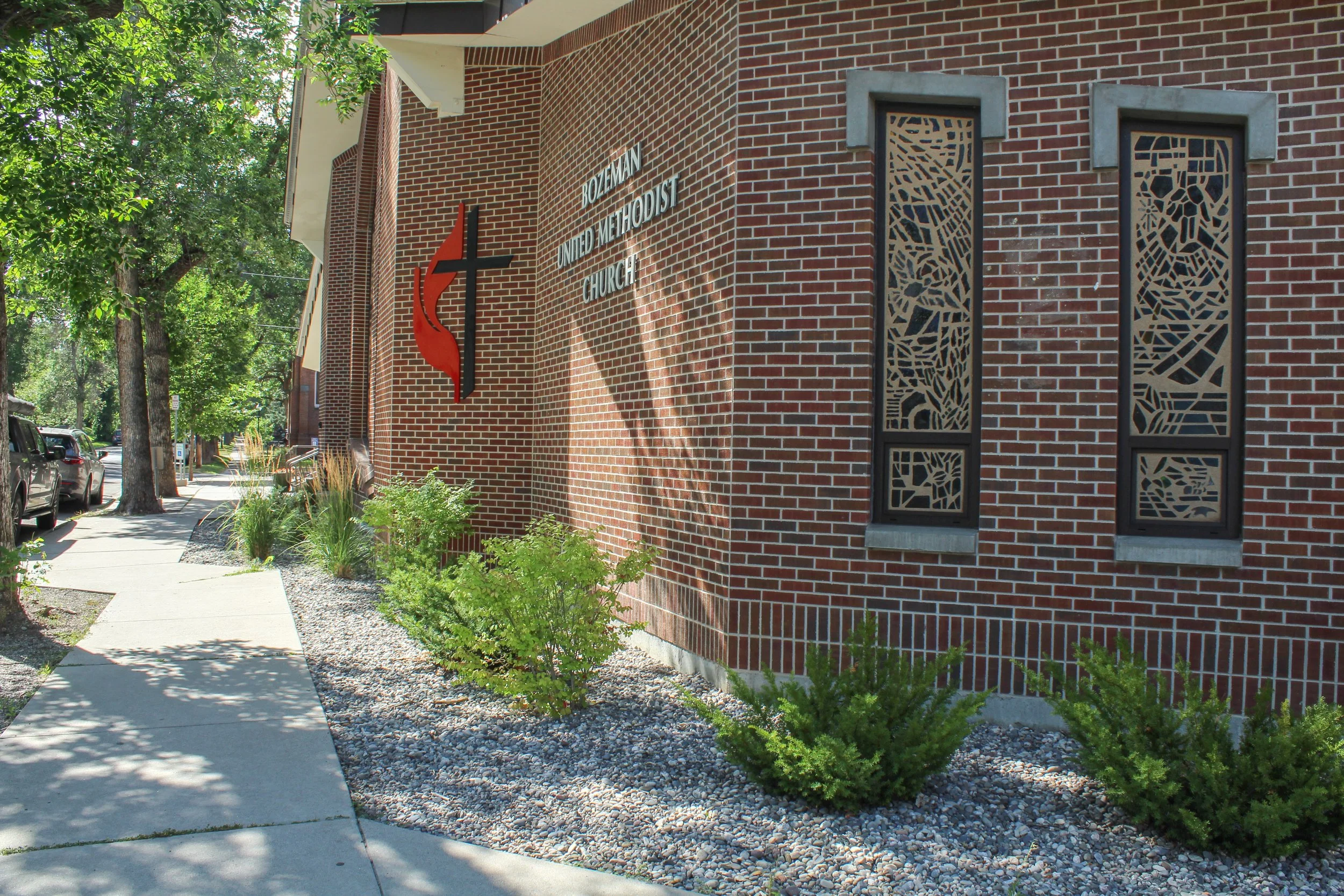 Exterior view of Bozeman United Methodist Church, a brick building with stained glass windows and church signage, sidewalk, parked cars, and greenery.