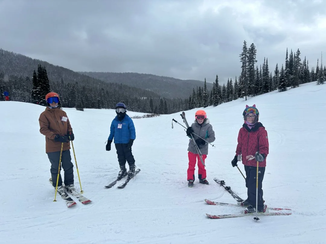 Four children in ski gear standing on a snowy mountain slope with trees and cloudy sky in the background.