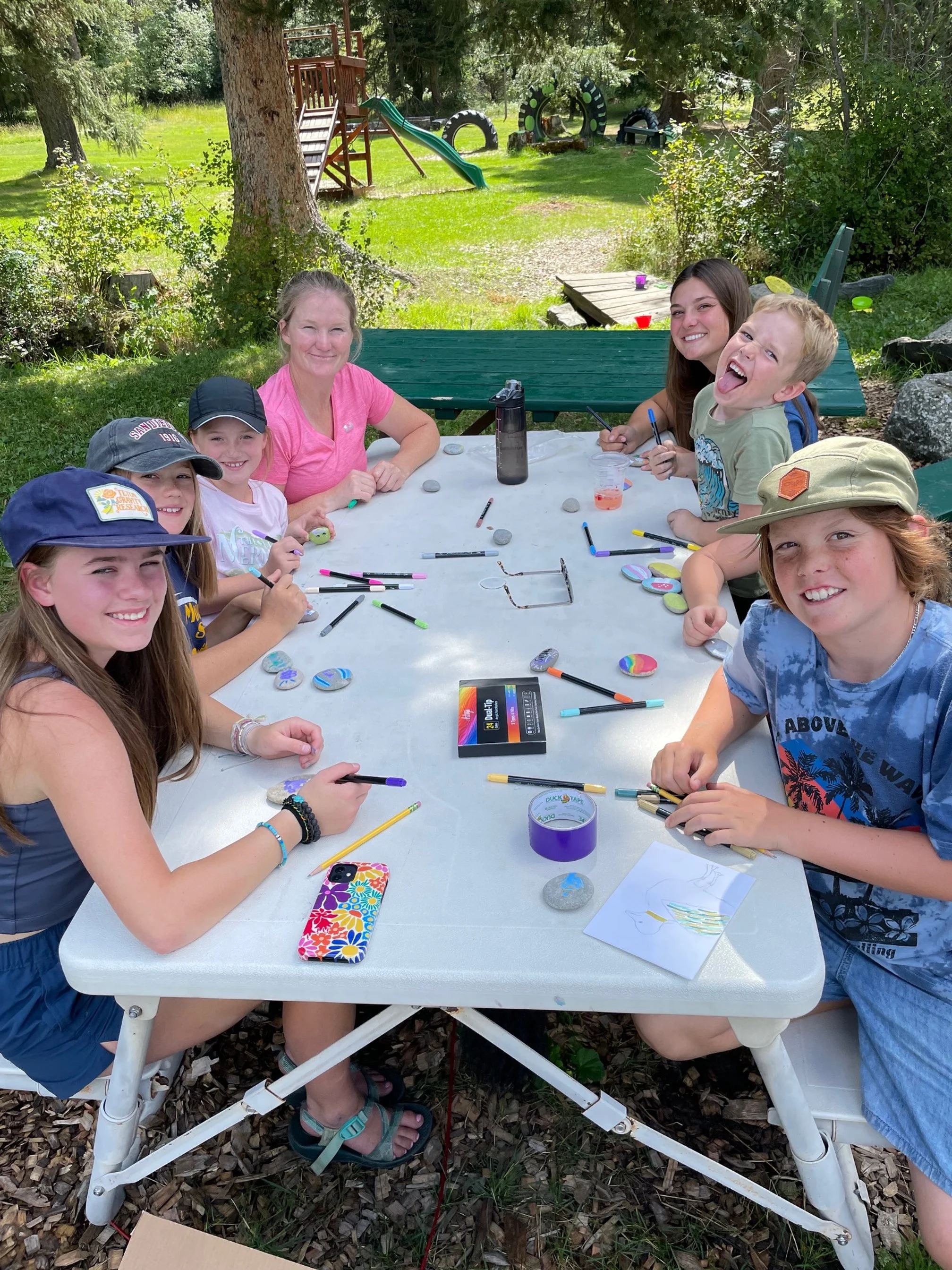 Group of children and a woman sitting at an outdoor table, engaged in arts and crafts, with markers and painted rocks on the table, in a backyard with trees, a playground, and a grassy area in the background.