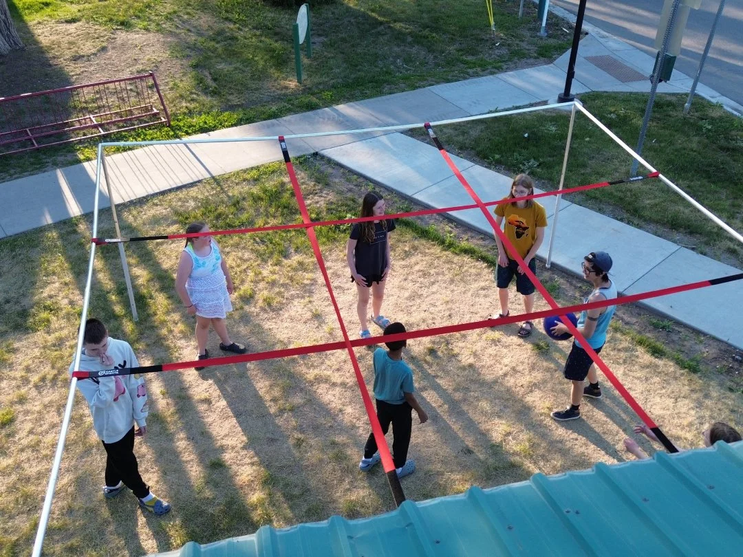 A group of children playing a volleyball game outdoors on a makeshift court with red and white lines marked on the ground, surrounded by grass and sidewalk. One child is preparing to serve while others are positioned on the court.