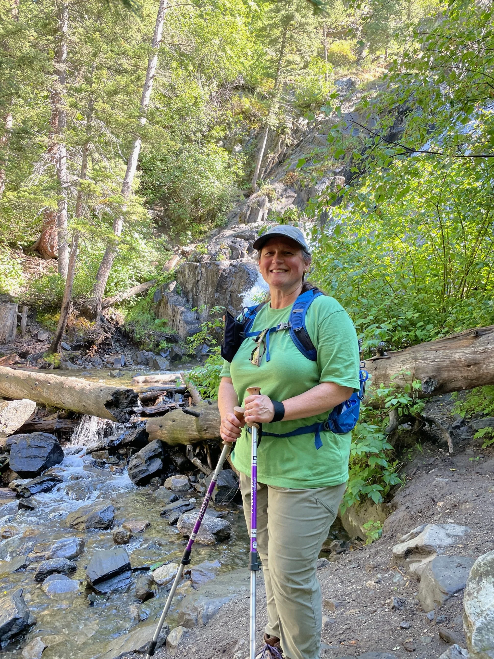 A woman with a backpack and hiking poles standing by a stream in a forest with trees and rocks in the background.
