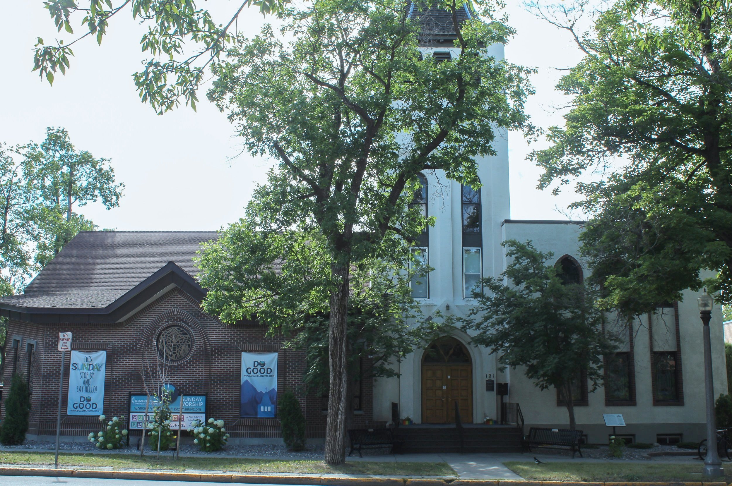 A church building with a tall steeple surrounded by trees, with banners on the front advertising Sunday worship and a do good message, and a sidewalk with benches and a lamp post.