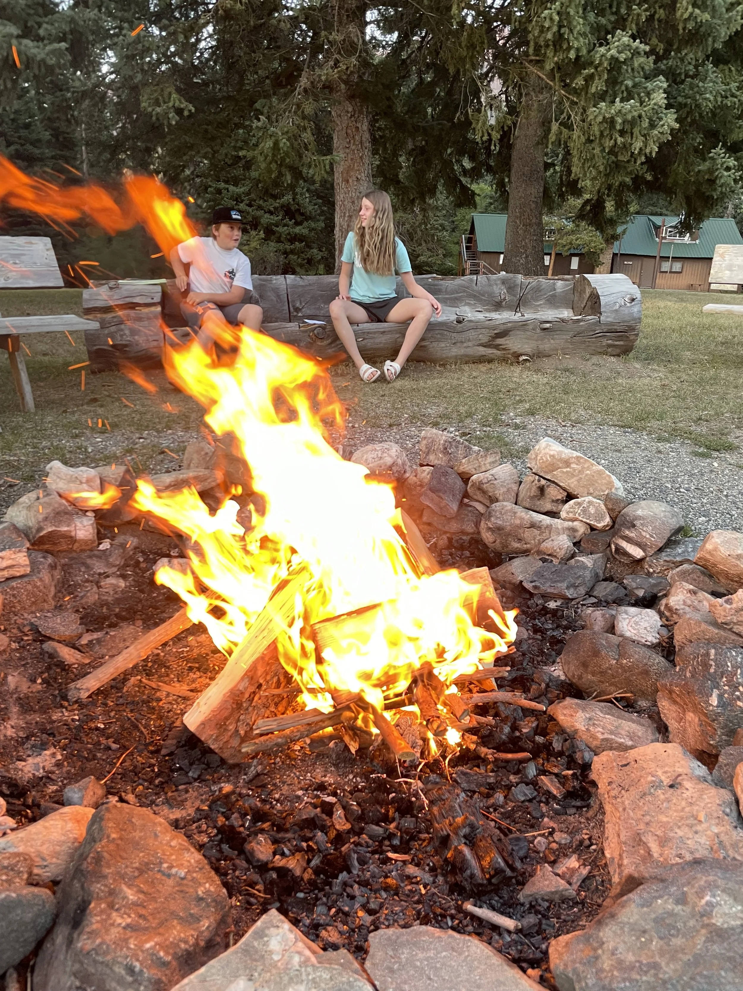 Two children sitting on a wooden log bench near a campfire outdoors with trees in the background.