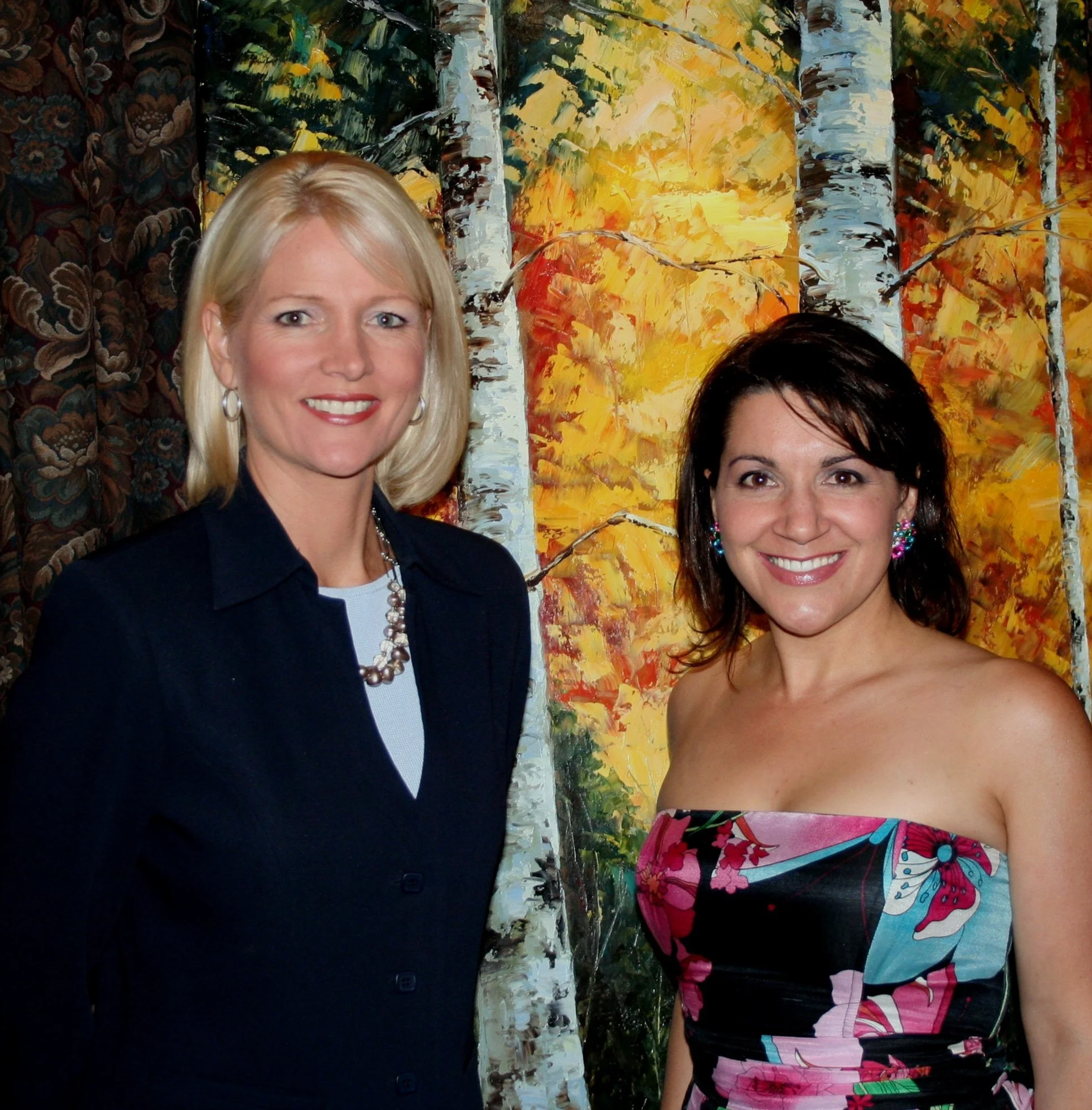 Two women at an art gallery or exhibit, with one woman with blonde hair and earrings smiling at the camera, and the other woman with dark hair and colorful earrings, standing in front of an abstract landscape painting of birch trees with vibrant yellow, orange, red, and green foliage.
