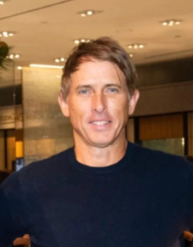 A man with short brown hair and blue eyes posing indoors with a smile, wearing a dark shirt, against a background with ceiling lights and brick walls.