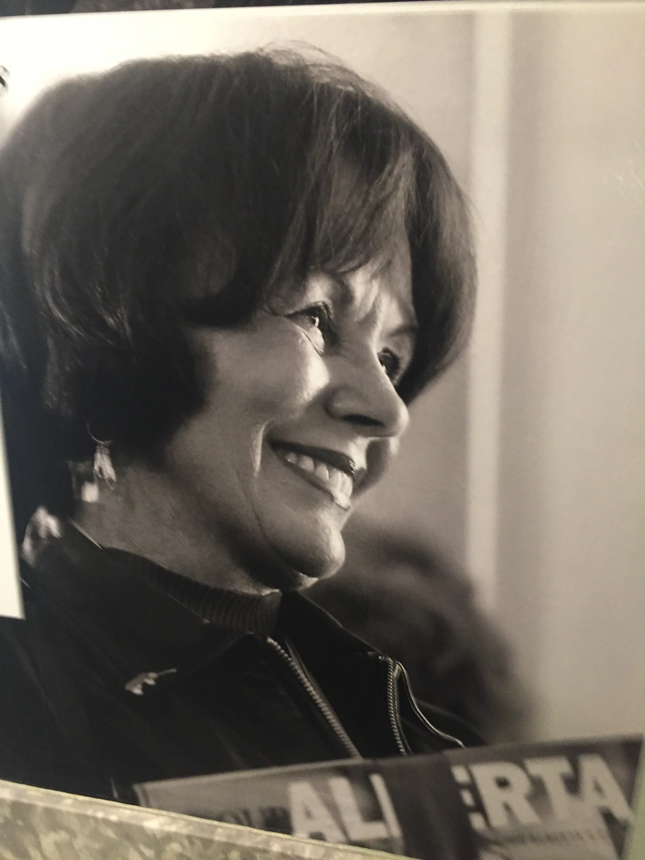 A woman with short dark hair, smiling, wearing earrings, and a jacket, captured in profile. The photo appears to be black and white.