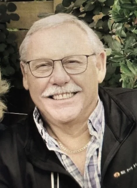 A smiling older man with glasses, white hair, and a white mustache, wearing a plaid shirt, a gold chain, and a black jacket, standing outdoors with green foliage in the background.