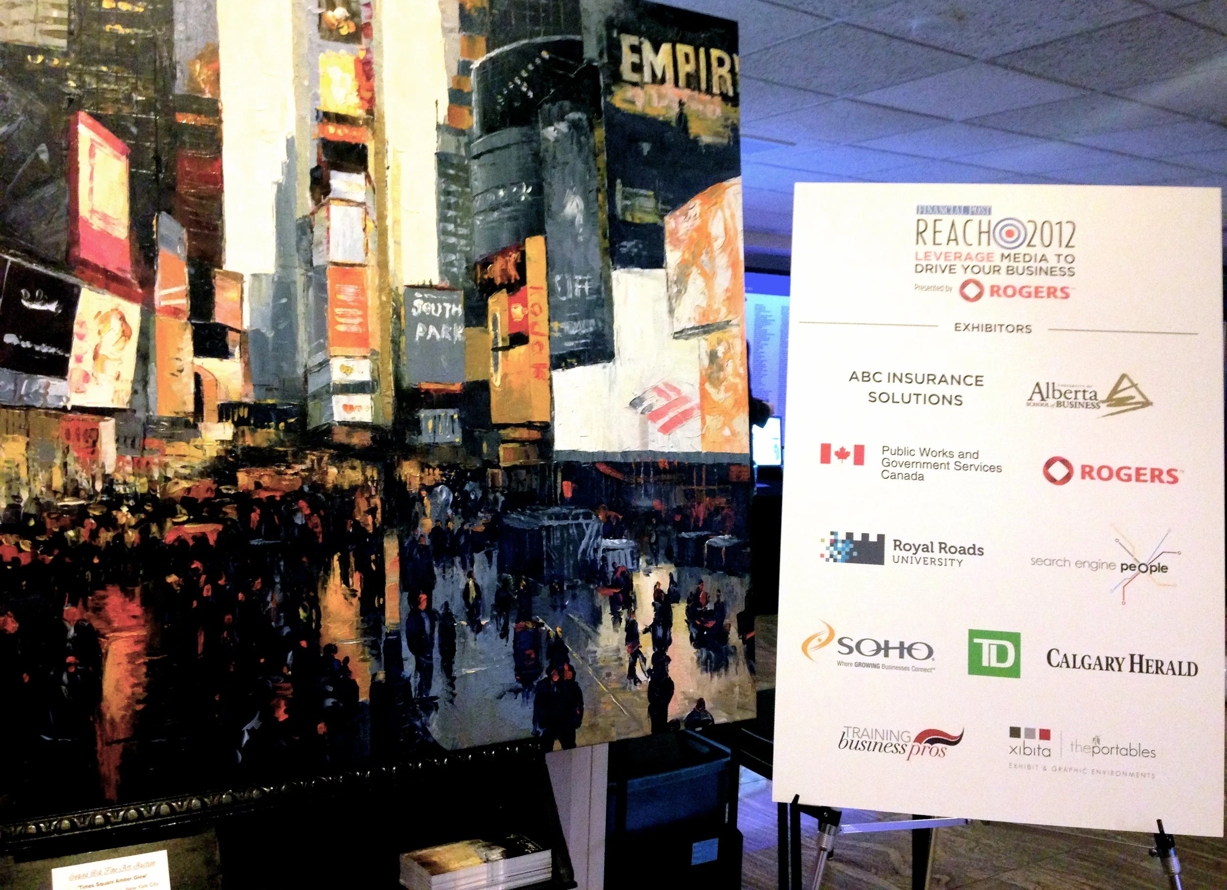 A colorful artistic cityscape painting depicting a busy street scene with illuminated buildings, crowd of people and reflections on wet pavement, displayed at an exhibition with promotional signage for various businesses.