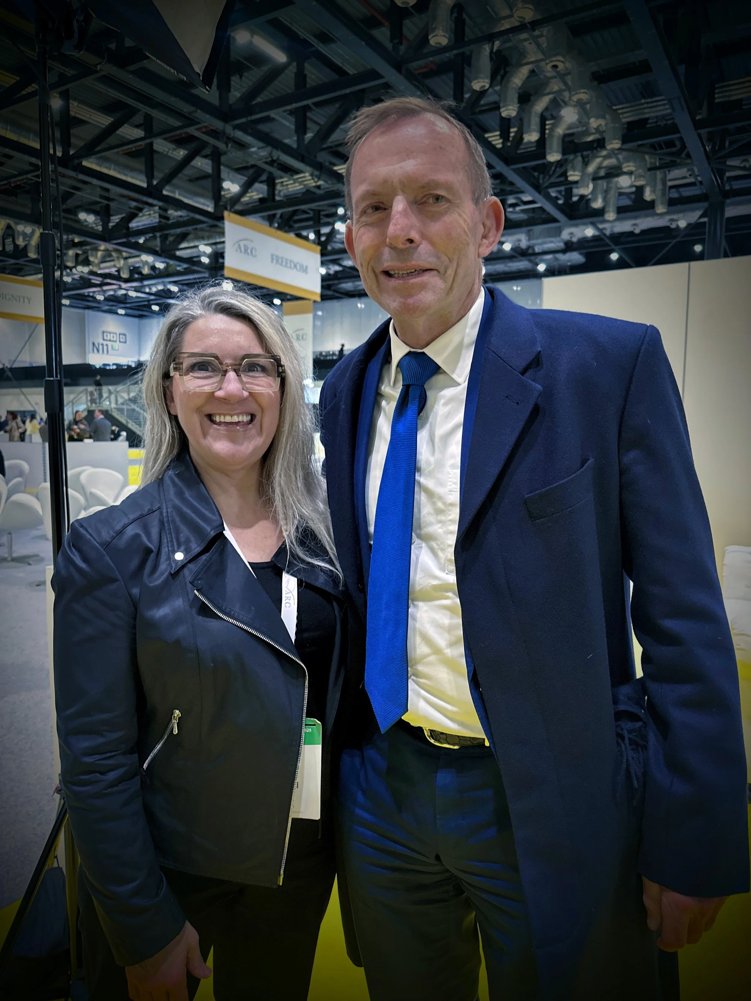 Two people, a woman with long blonde hair and glasses wearing a black leather jacket, and a man with short hair in a suit, are smiling and standing close together at an indoor event or conference, with signs and other attendees visible in the background.