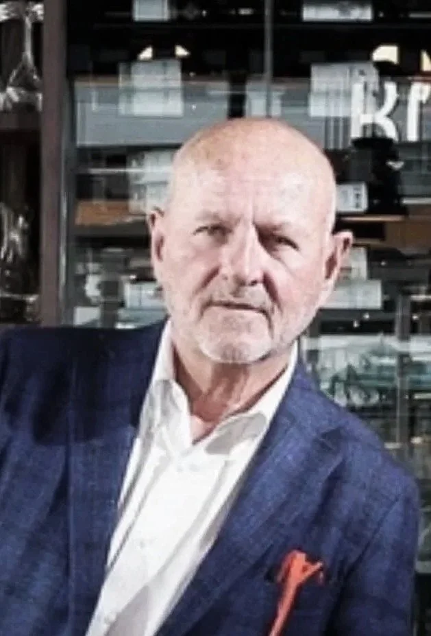 An older man with a serious expression, light-colored blazer, white shirt, and a red pocket square, standing in front of a glass storefront with shelves inside.