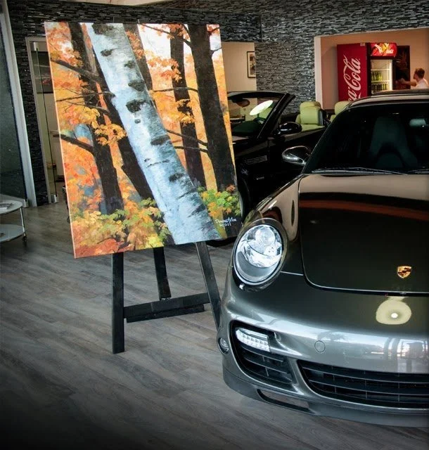 A black Porsche sports car parked indoors next to an easel holding a painting of a forest with orange and yellow autumn leaves and white-barked trees.